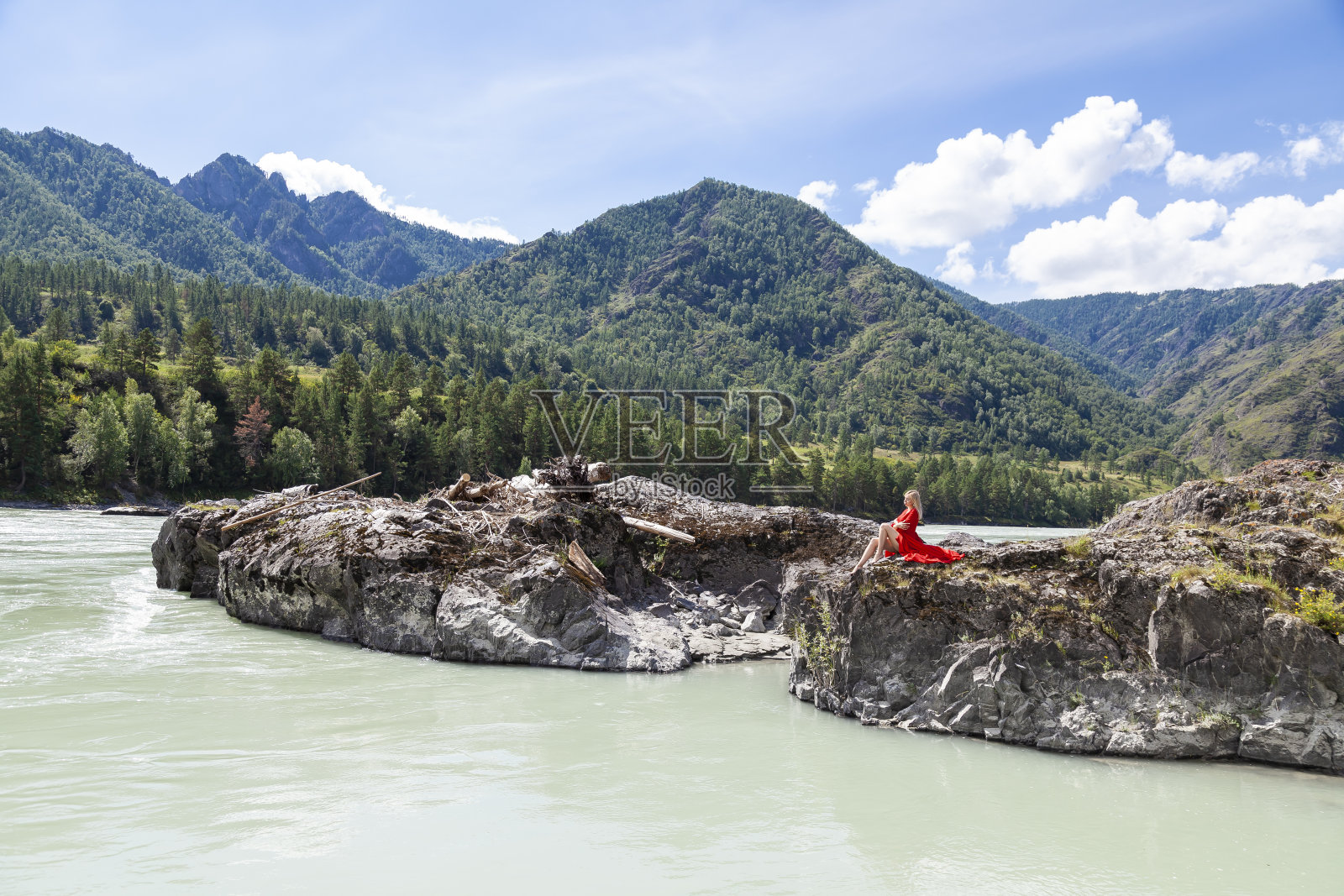 身穿红色复古连衣裙的金发女子，双臂交叉放在胸前，拥抱着自己，坐在卡顿河岸边的岩石上伸展她的长腿，背景是夏日的群山。照片摄影图片