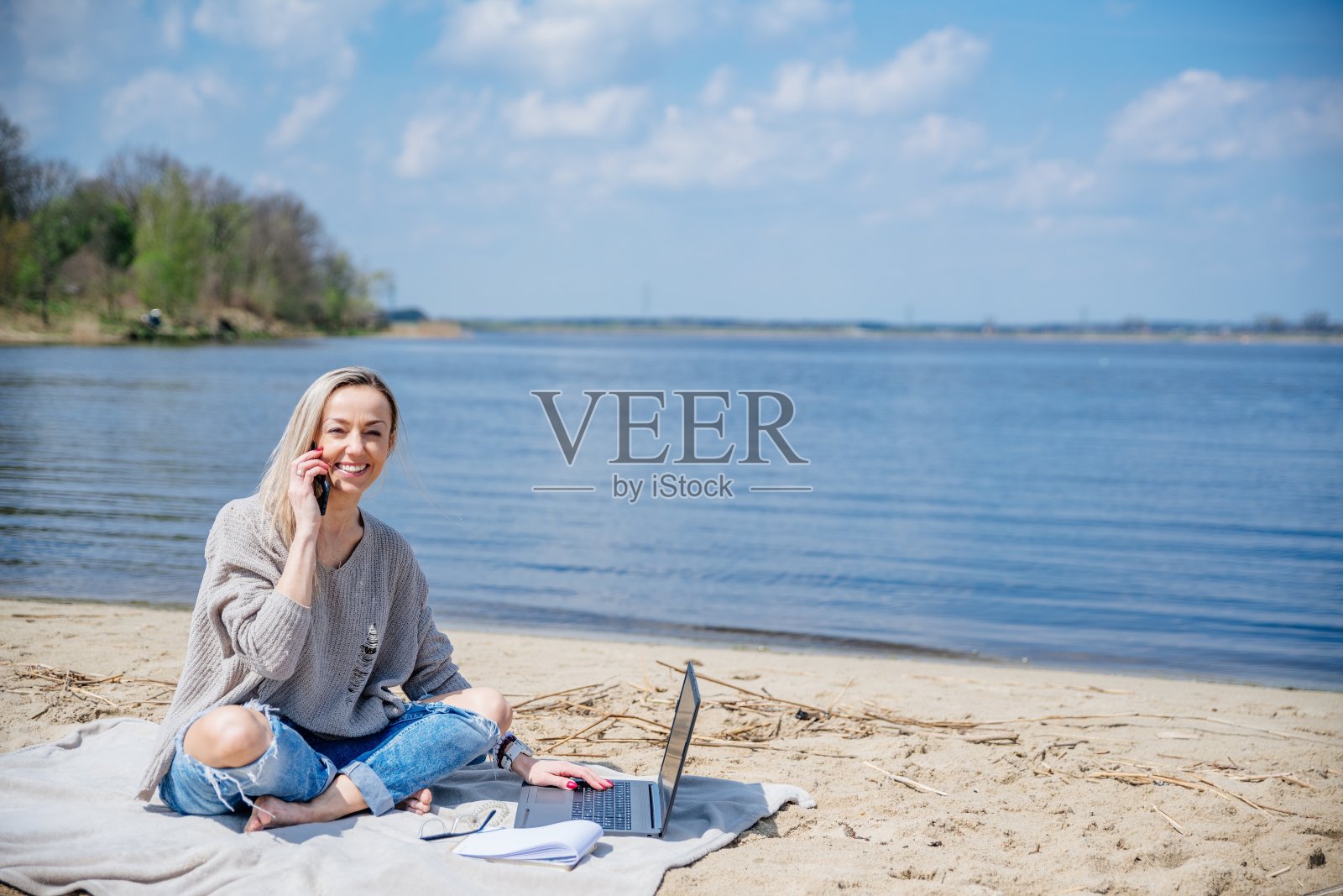 一个阳光明媚的日子，微笑的金发女郎在湖边远程工作。照片摄影图片