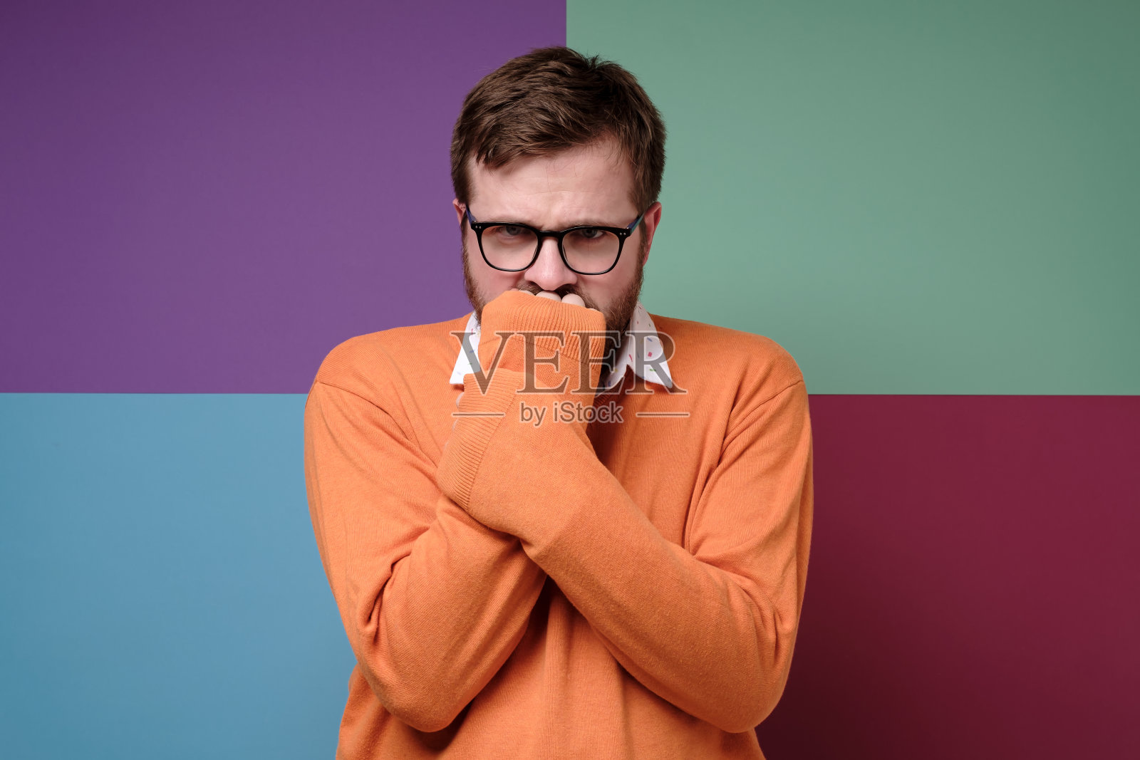 沮丧的男子皱着眉头，把拳头藏在毛衣的袖子里，用手捂住嘴，愤怒地看着摄像机。照片摄影图片