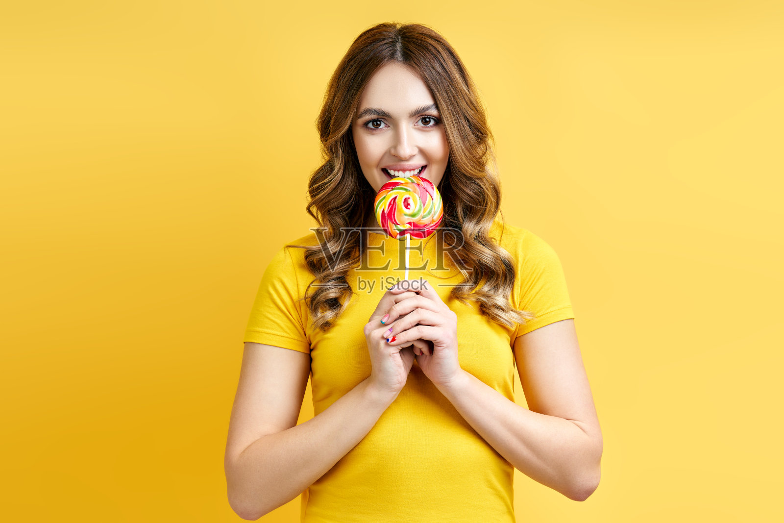 年轻女子咬美味的彩色棒棒糖黄色的背景照片摄影图片