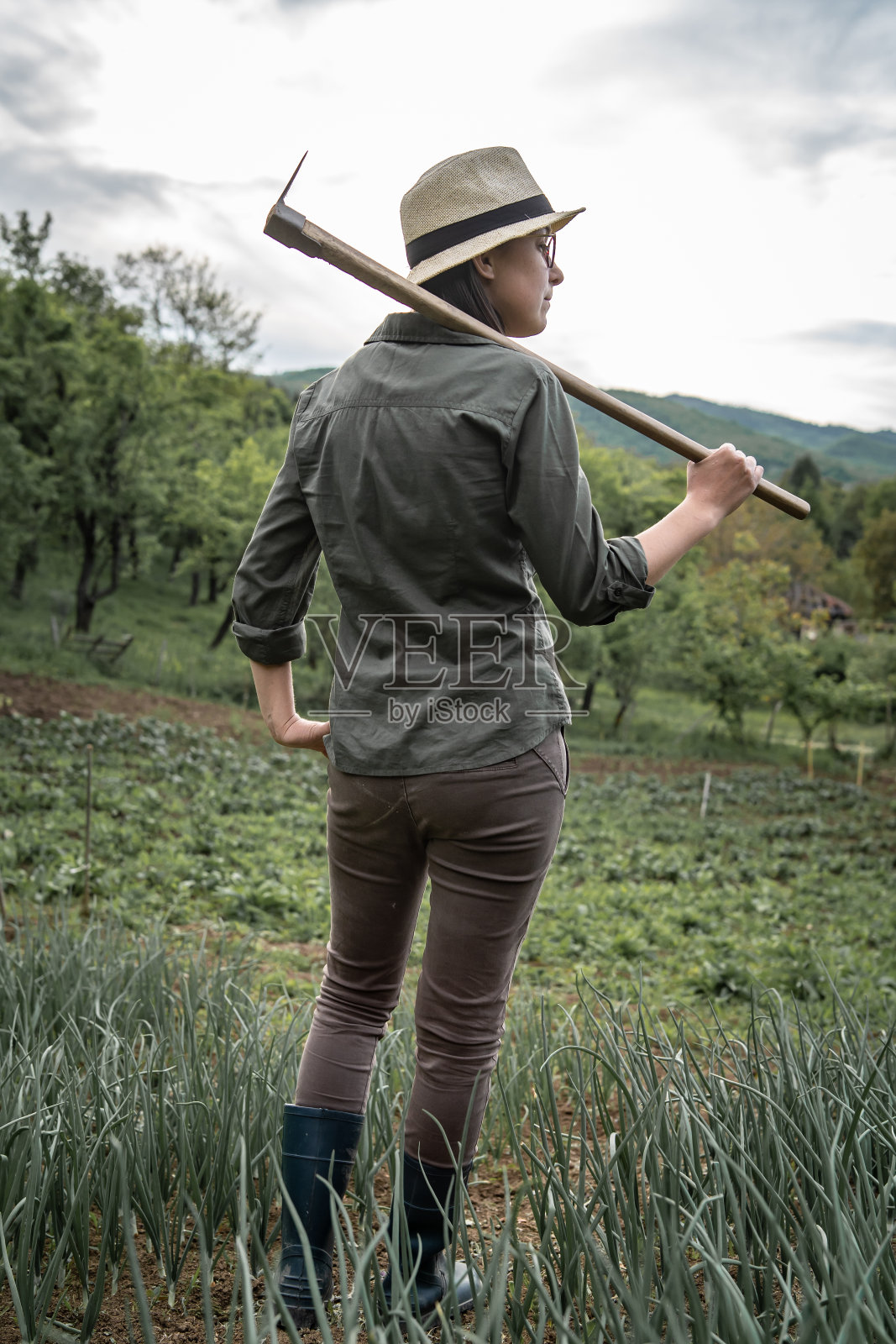 年轻的成年白人妇女女性农民的背影肖像站在农田准备植物与锄头在她的肩膀上摆姿势在夏天看向一边照片摄影图片