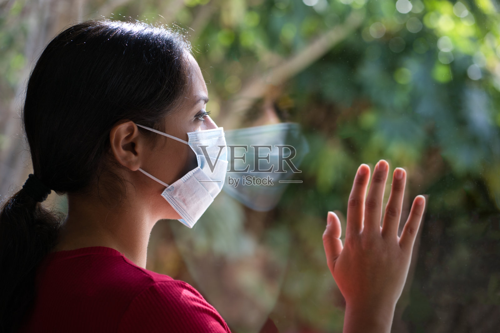 一个戴着面罩的黑发女人透过窗户看着玻璃上的倒影，在家里自我隔离。照片摄影图片