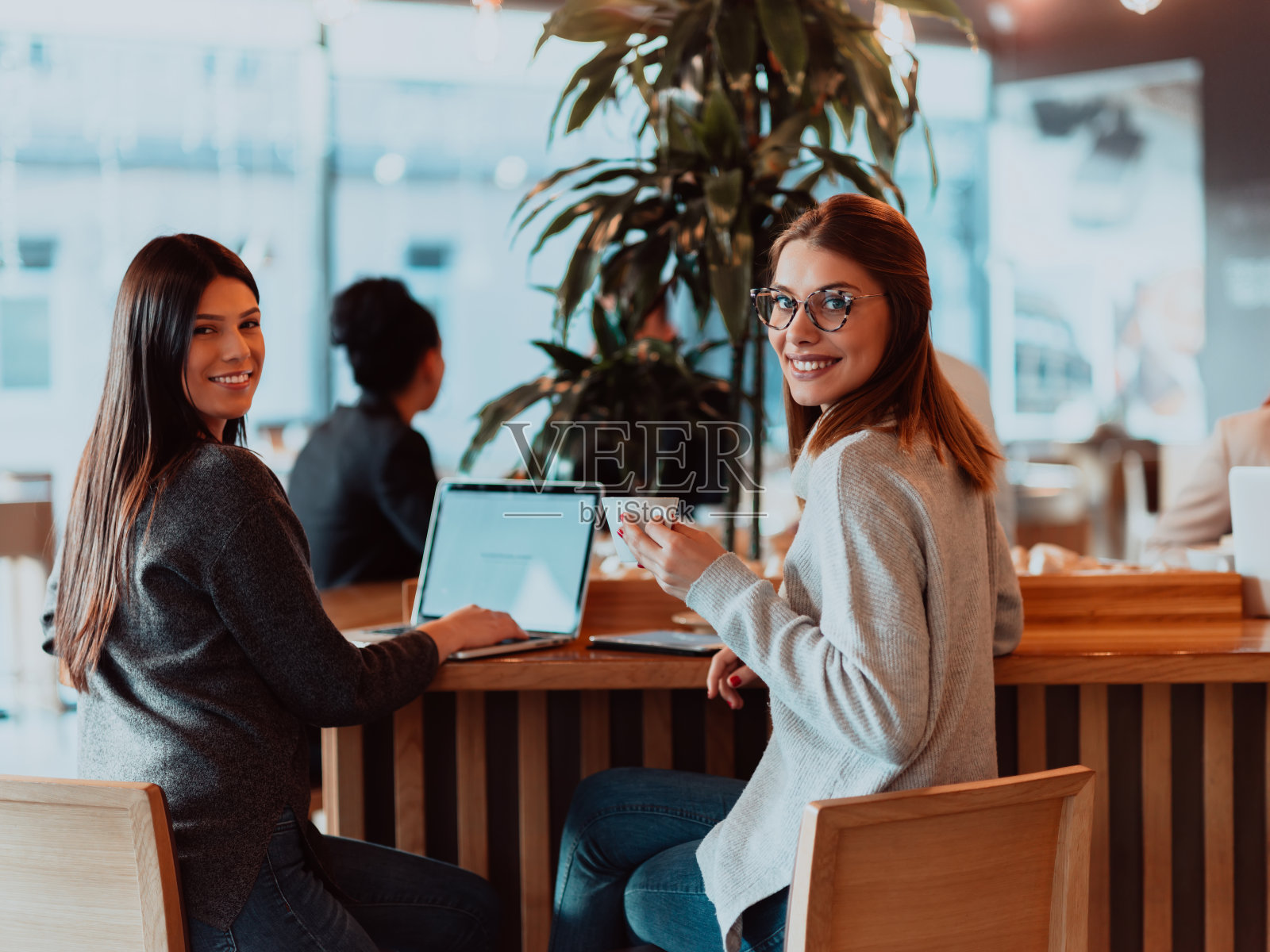 两个年轻的女商人坐在咖啡馆的桌子旁。女孩在笔记本电脑屏幕上显示同事信息。女孩用智能手机，写博客。团队合作、商务会议. .照片摄影图片
