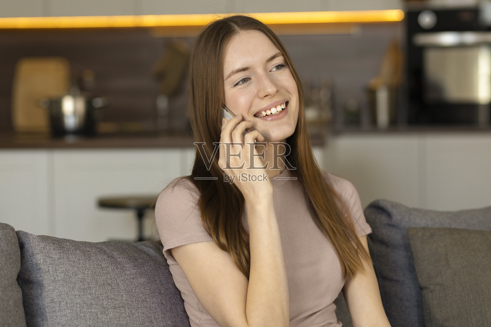 快乐的千禧一代白人女性在客厅里打电话。快乐的女孩坐在家里的沙发上愉快地打电话照片摄影图片