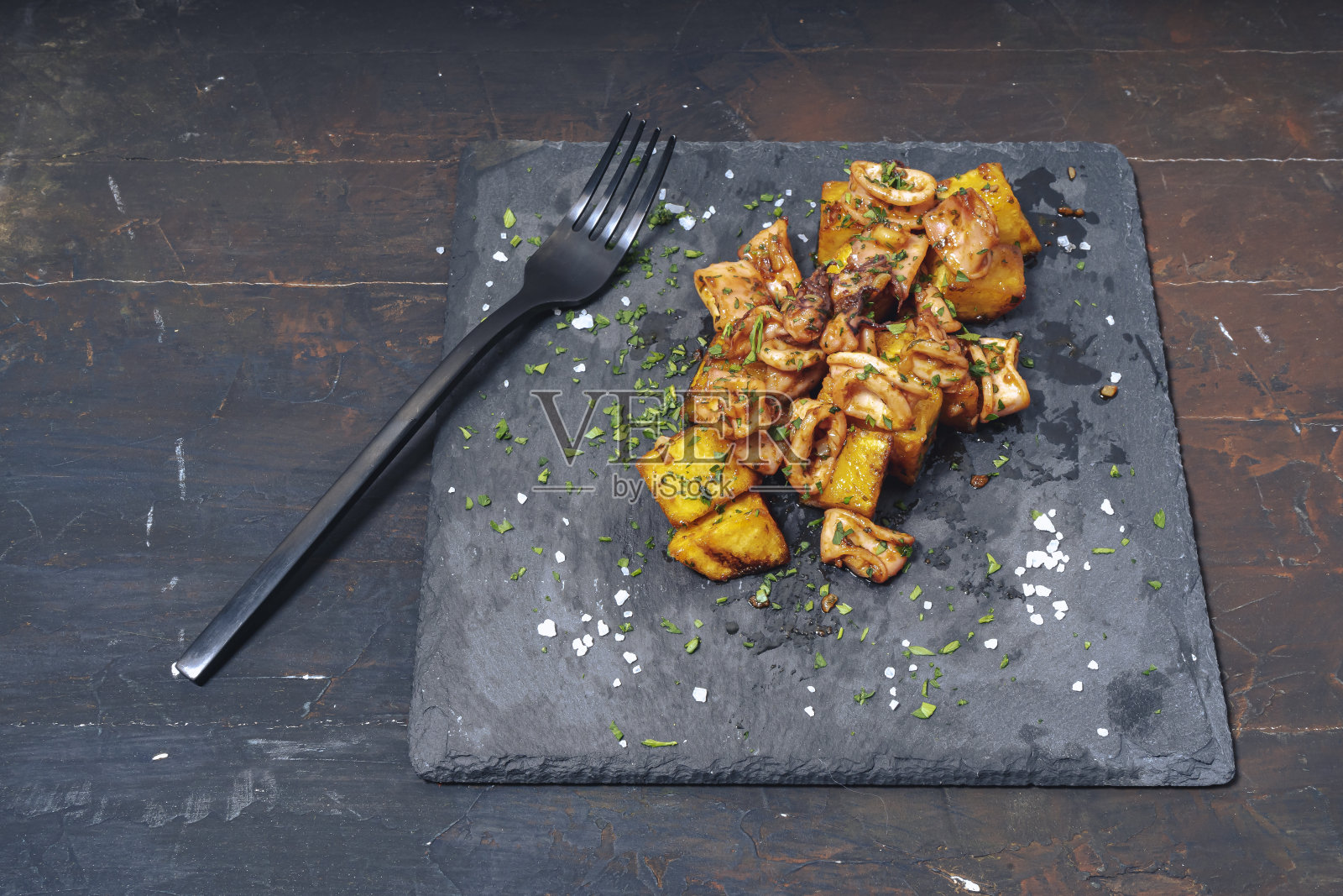 西班牙的小鱿鱼或花栗鼠配大蒜和土豆，这是典型的地中海西班牙菜的开胃菜，以美食的方式放在黑色的板上，用叉子品尝。传统美食的概念。照片摄影图片