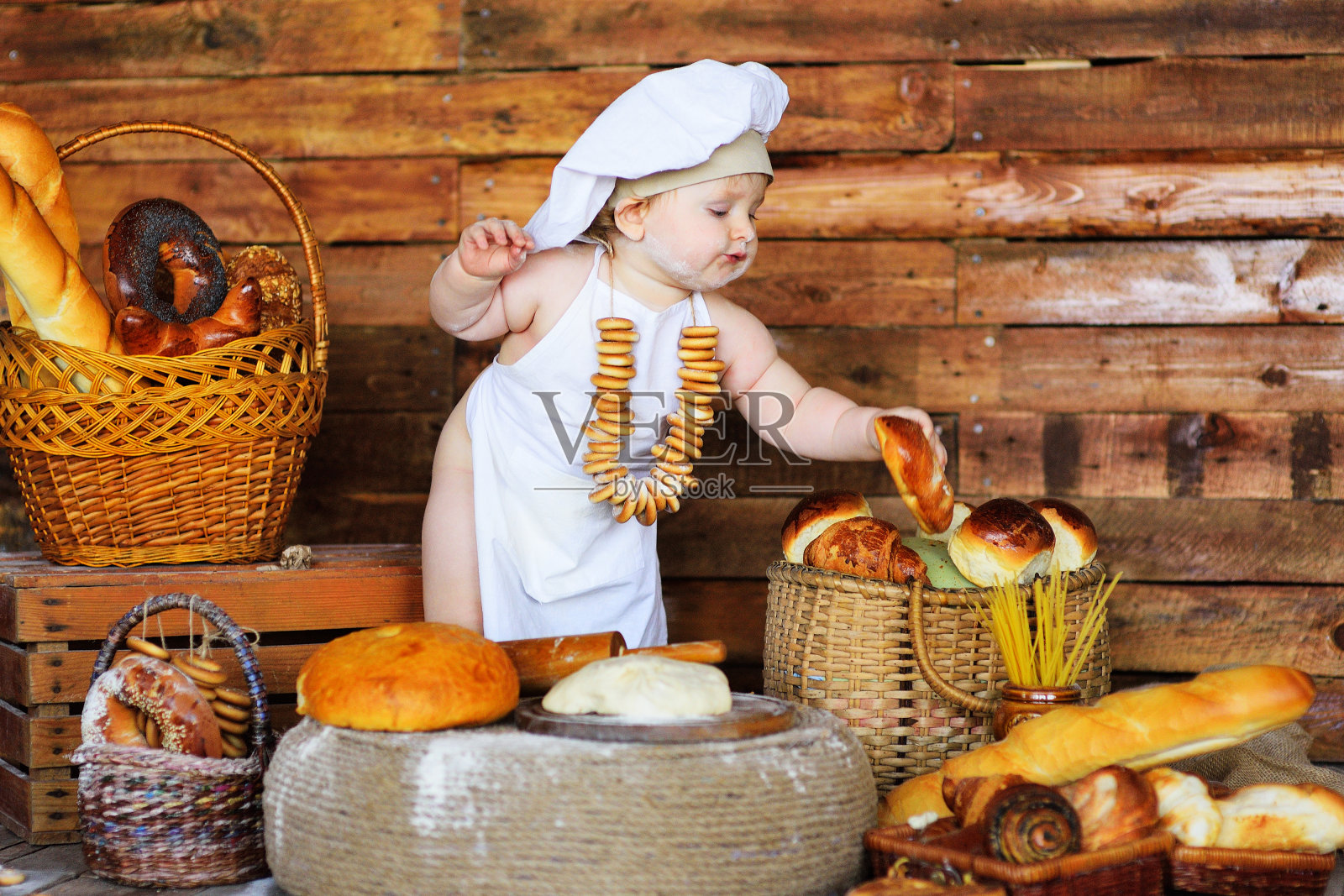 小男孩戴着厨师帽，系着围裙，脖子上围着一串百吉饼，坐在一家面包店里的面包房里照片摄影图片