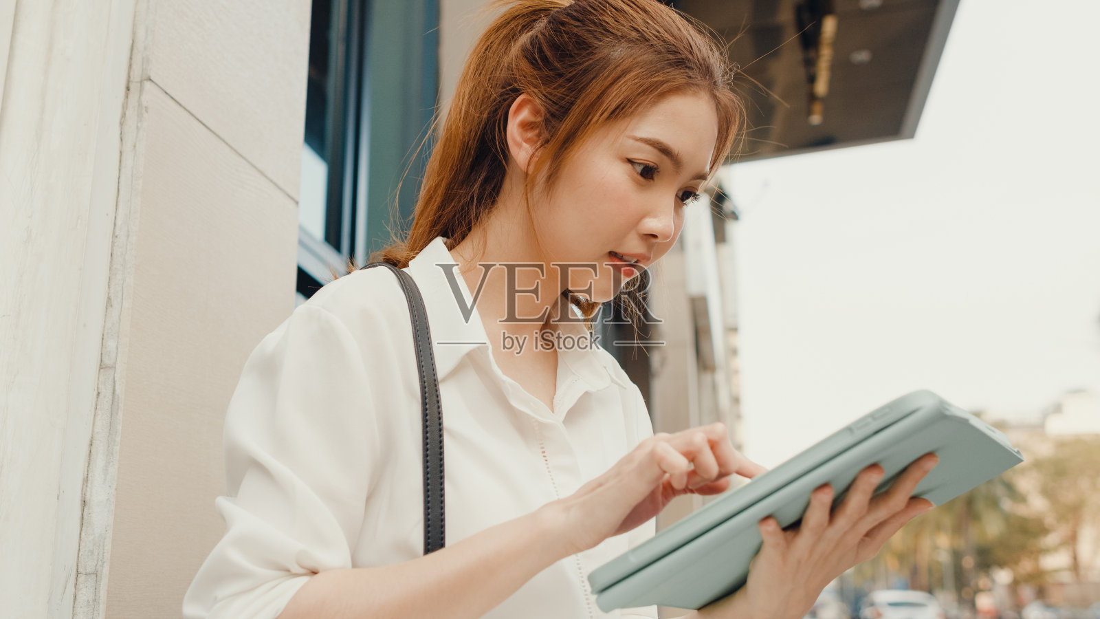 在现代都市中，年轻的亚洲成功女商人穿着时尚的办公服装，独自坐在户外，用数码平板电脑打字。照片摄影图片