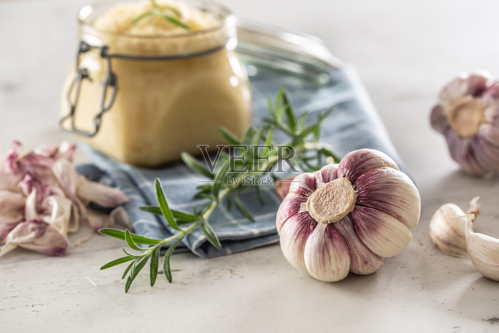 自家种植的大蒜灯泡放在一个装满新鲜大蒜酱的玻璃瓶前，铺在铺有迷迭香的厨房布上。照片摄影图片