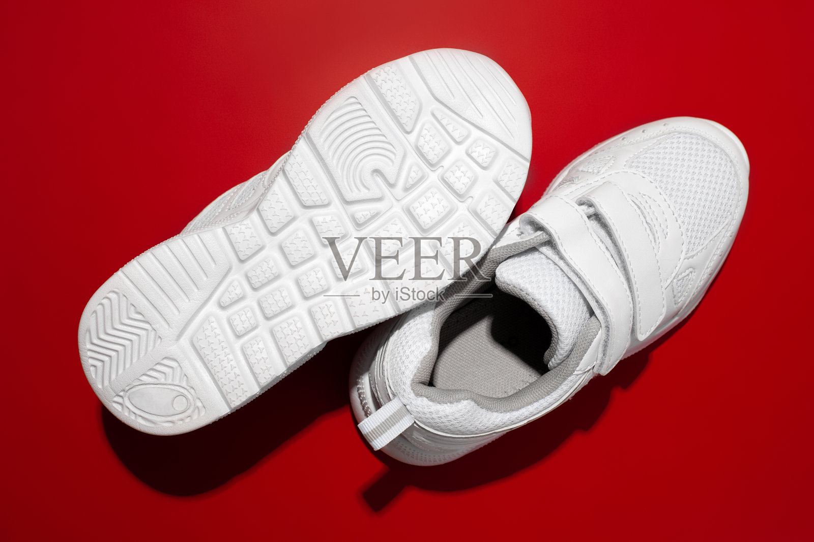 顶视图两个白色的小孩运动鞋与魔术贴带与红色背景上的太阳阴影，一个运动鞋躺在鞋底上照片摄影图片