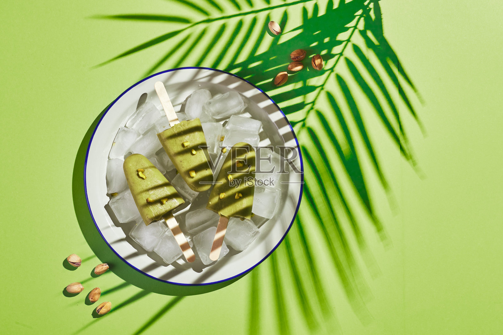 冰冻自制开心果冰棒在碗冰上的绿色背景与棕榈叶阴影在强光下。清凉的冰棒，冰冻的绿汁棒。俯视图，复制空间照片摄影图片