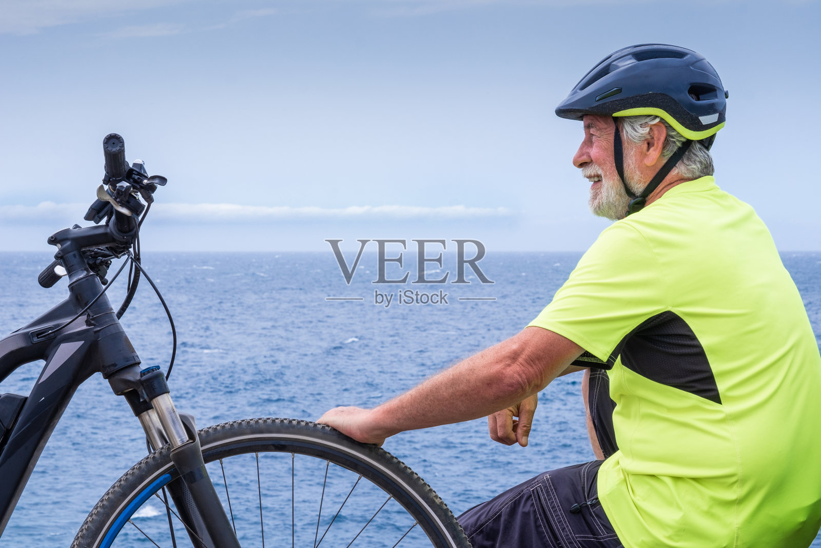 微笑的高级自行车男子休息在海边的悬崖远足后与他的电动自行车。照片摄影图片