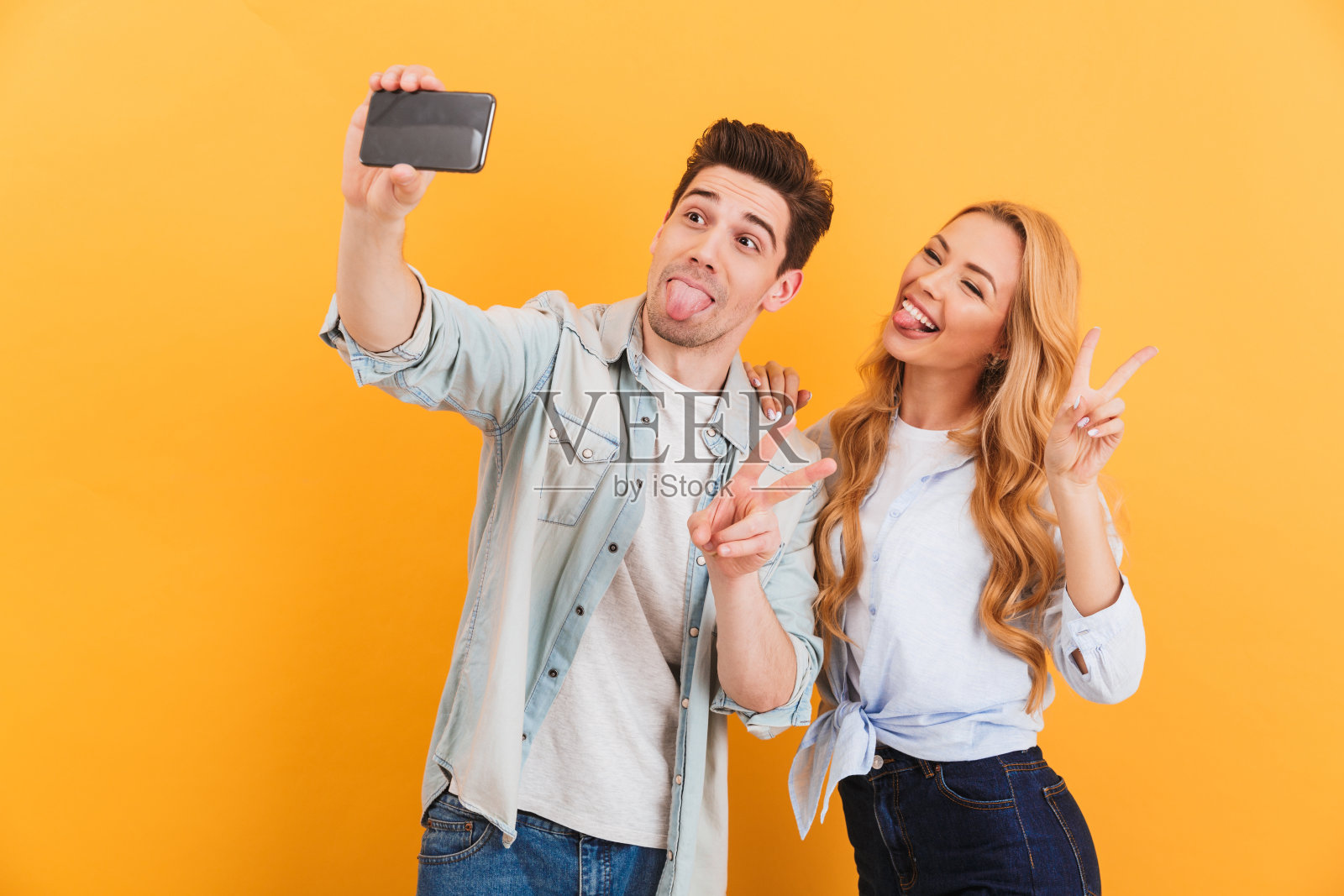 两个有趣的男人和女人在智能手机上自拍，同时显示胜利标志，孤立在黄色背景照片摄影图片