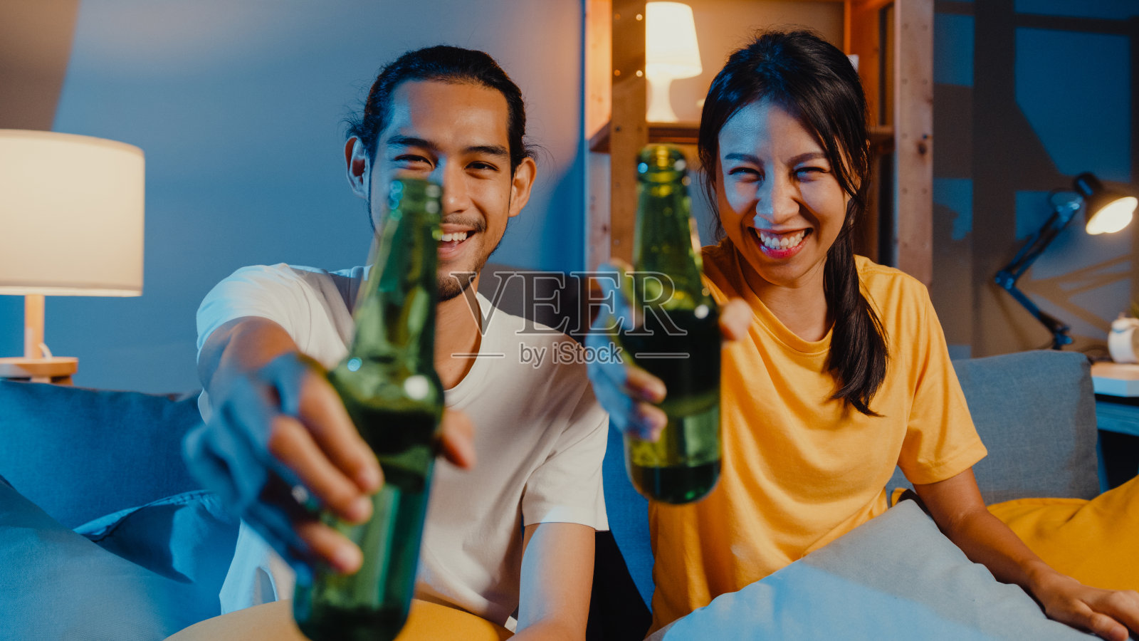 快乐的年轻亚洲夫妇看着摄像机享受夜晚聚会活动在线坐在沙发上与朋友视频通话，通过视频通话在家里的客厅喝啤酒。照片摄影图片