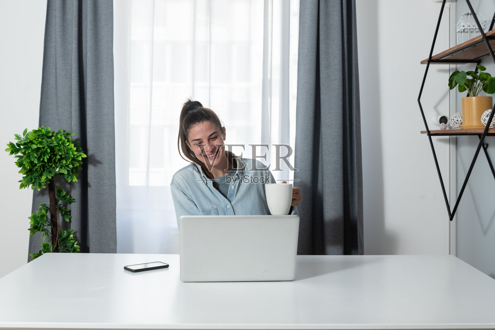 年轻快乐满意的自由职业者商务女性从工作中休息在家的家庭办公室和喝咖啡从杯子聊天与她的同事在线视频电话照片摄影图片