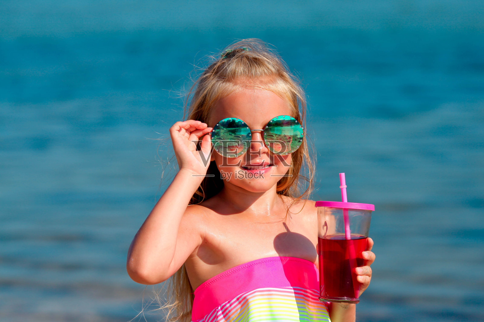 漂亮的小女孩穿着泳衣，戴着太阳镜站在海边，手里拿着一杯水果饮料。本空间照片摄影图片