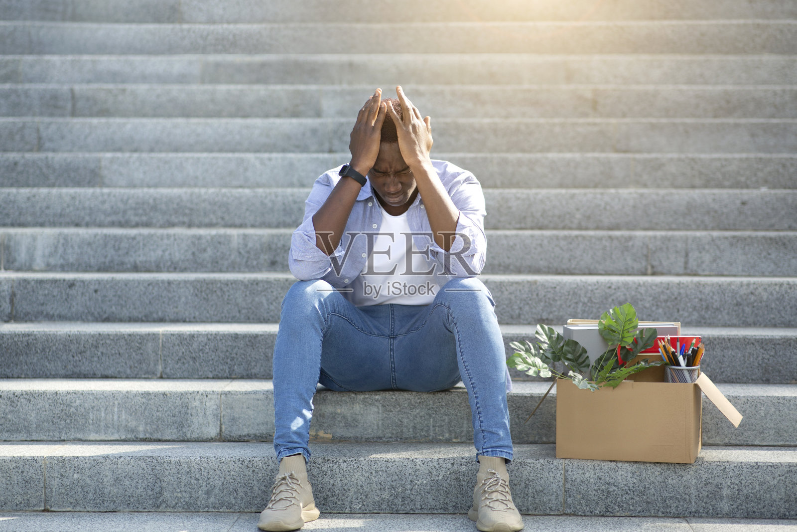 经济危机和破产。一个失业后拿着私人物品坐在楼梯上的绝望黑人，在外面照片摄影图片