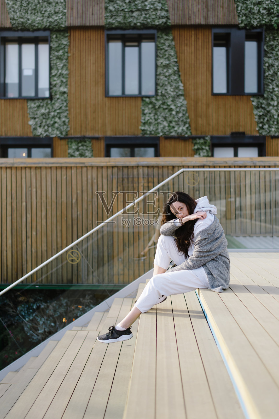 漂亮可爱的年轻女人穿着超大号帽衫坐在木台阶上。现代的观点和背后的建筑照片摄影图片