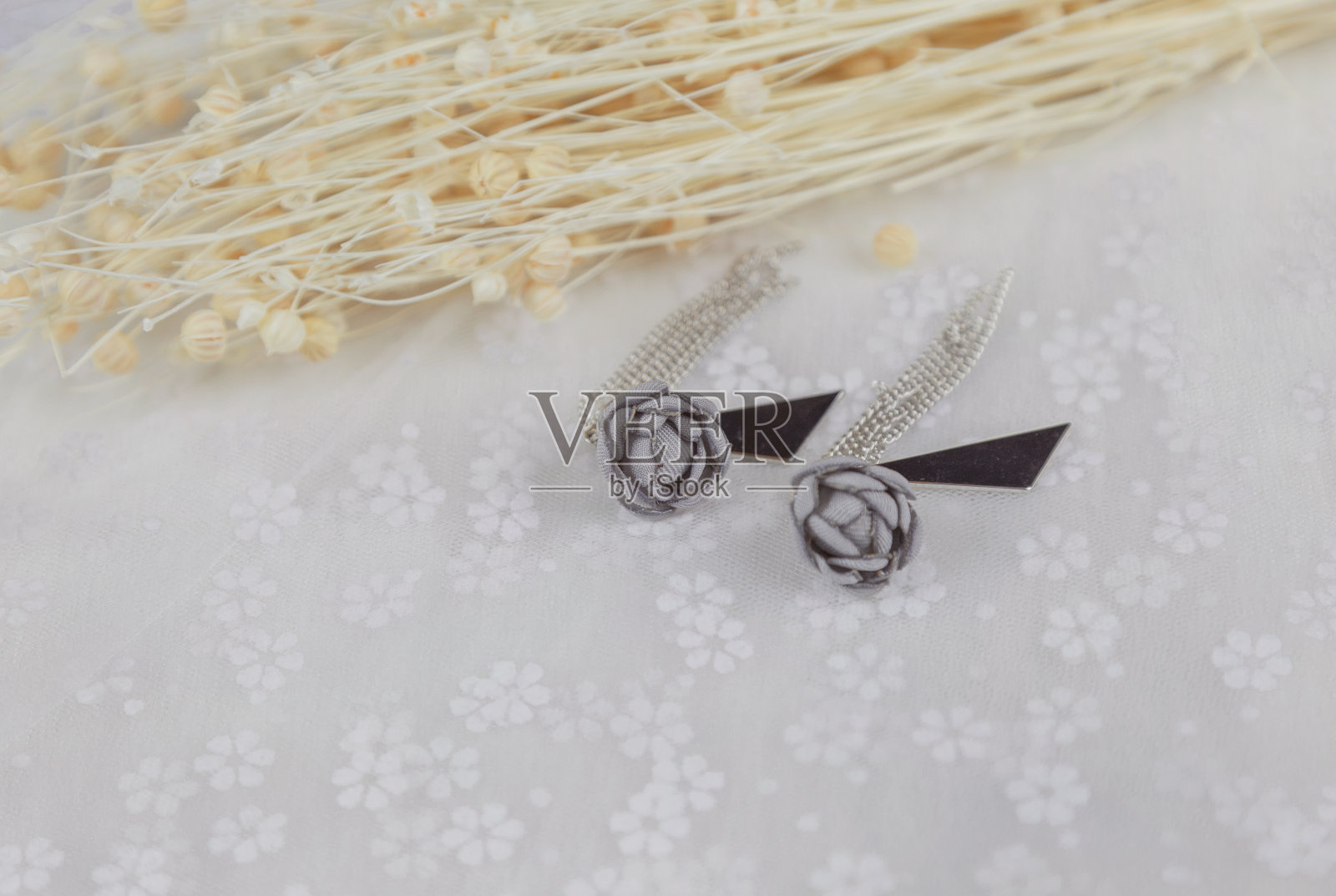 灰色吊坠耳环，它的形状是一朵花。在耳环旁边放一束花的标本。这些东西都放在薄纱上。这对耳环看起来非常优雅时尚。耳环以流苏和金属三角形装饰。照片摄影图片