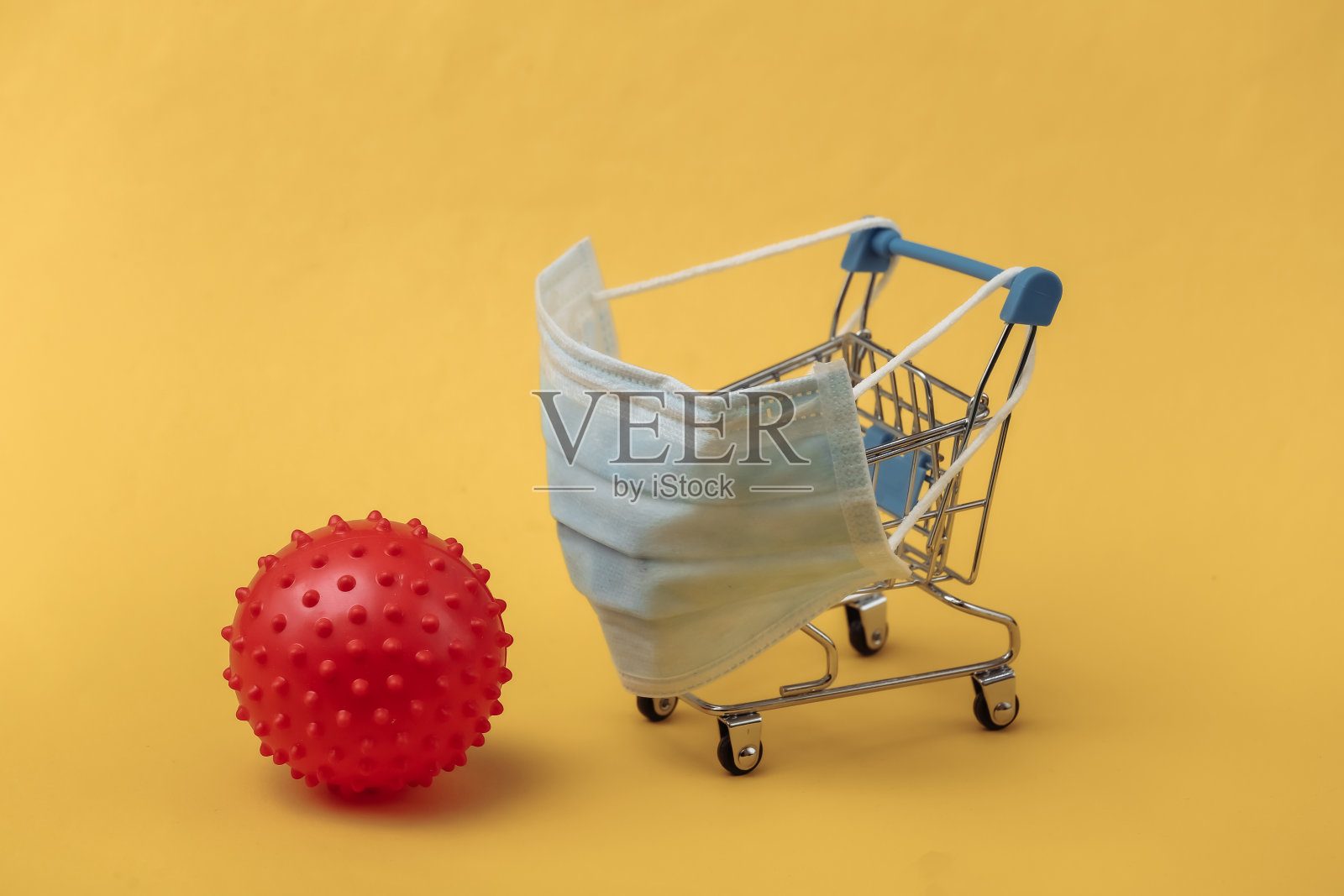装有医用口罩的迷你超市手推车，黄色背景上有病毒株模型。在新冠肺炎大流行期间购物照片摄影图片