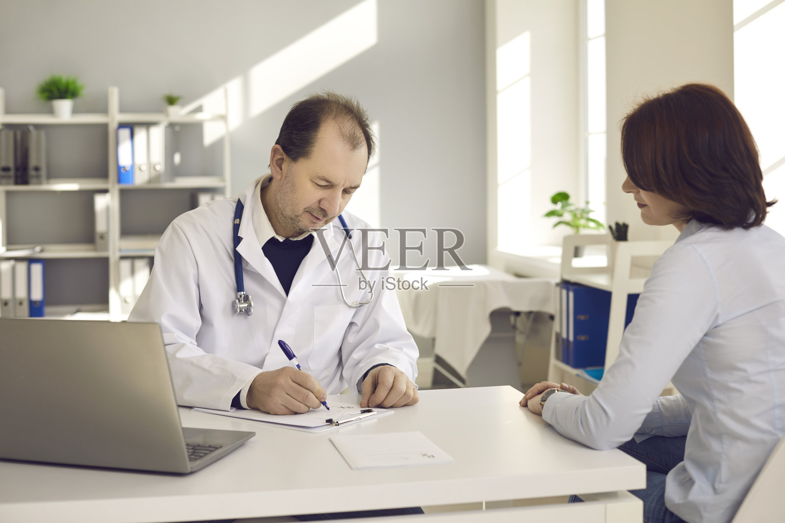 中年医生在他的医院办公室里给一位女病人开处方。照片摄影图片