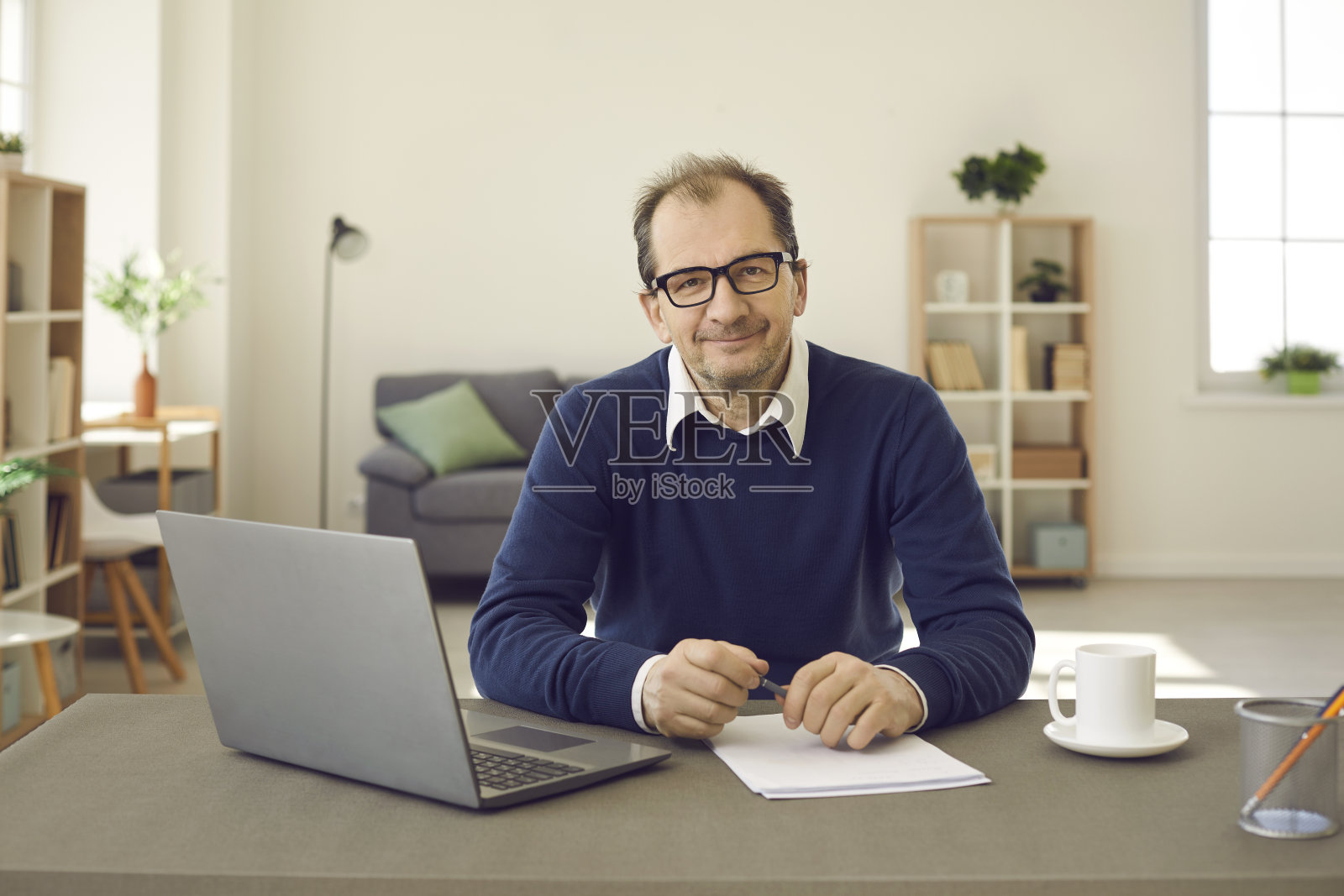 一个快乐的中年男人，戴着眼镜，坐在家庭办公室的桌子前对着镜头微笑照片摄影图片