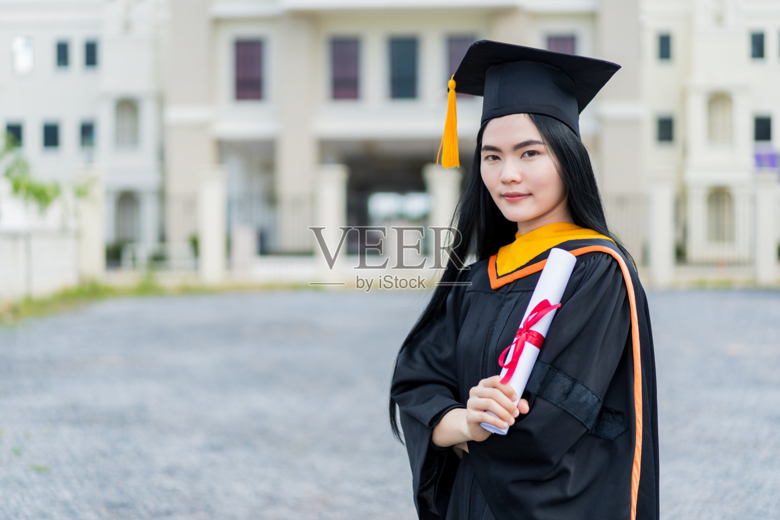 一位年轻漂亮的亚洲女大学毕业生穿着毕业礼服和学士帽，拿着学位证书站在大学楼前参加毕业典礼照片摄影图片