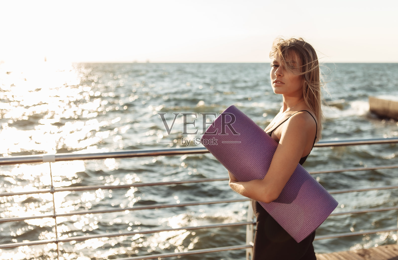 年轻苗条健康的女人在日出前在海滩上抱着瑜伽垫。照片摄影图片