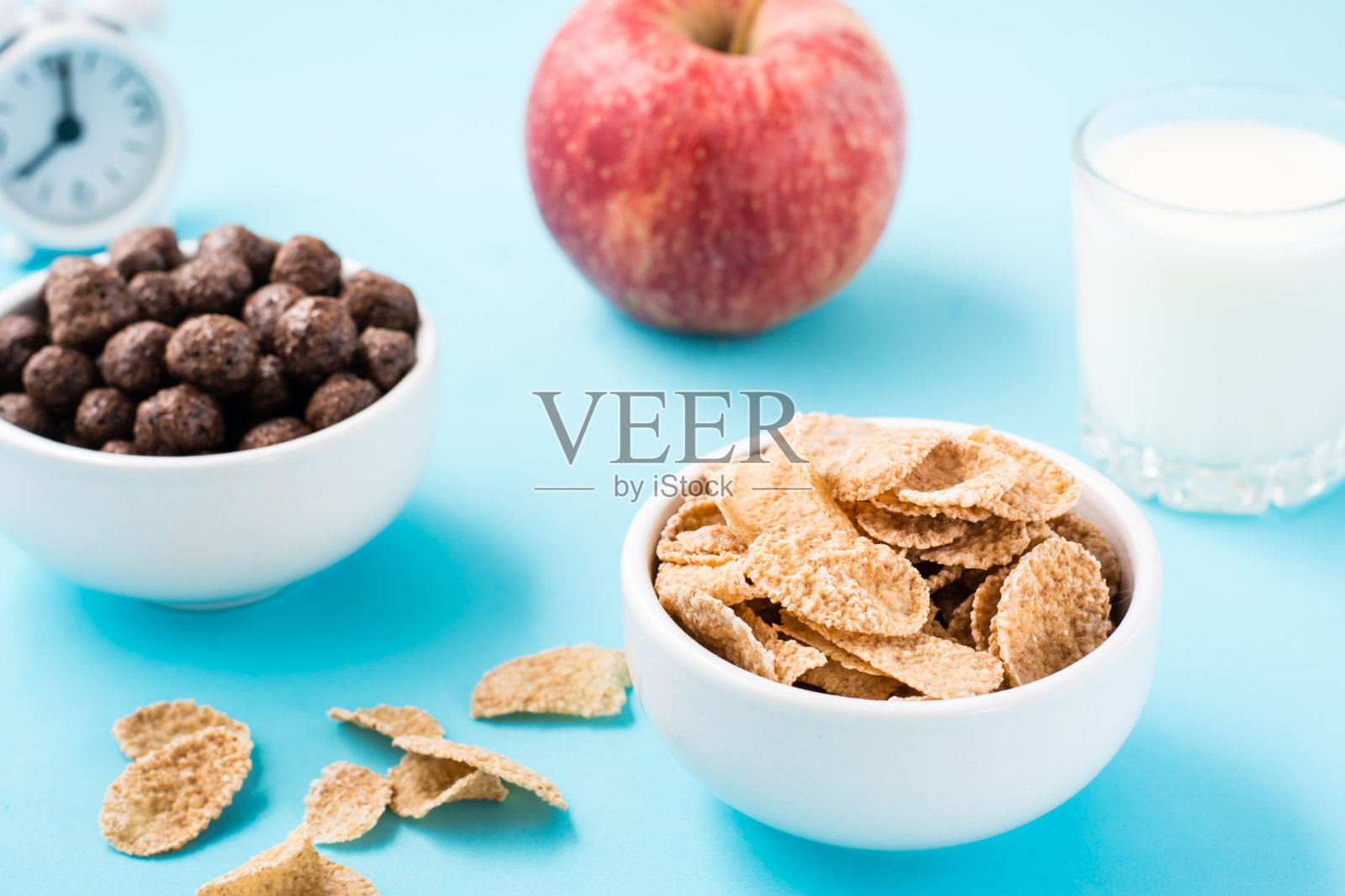 有麦片和巧克力球的碗，一杯牛奶，一个苹果和一个蓝色背景的闹钟。安排早餐。特写镜头照片摄影图片