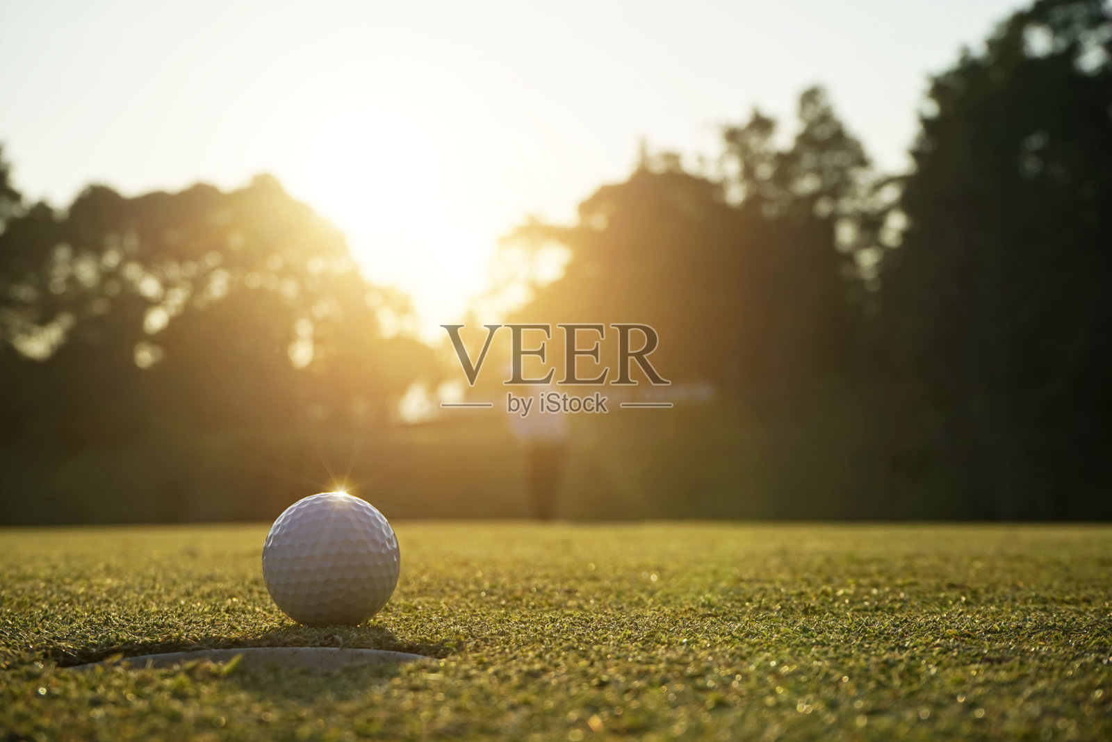 高尔夫球手把高尔夫球在绿色的高尔夫上，镜头光晕在日落黄昏时间。照片摄影图片