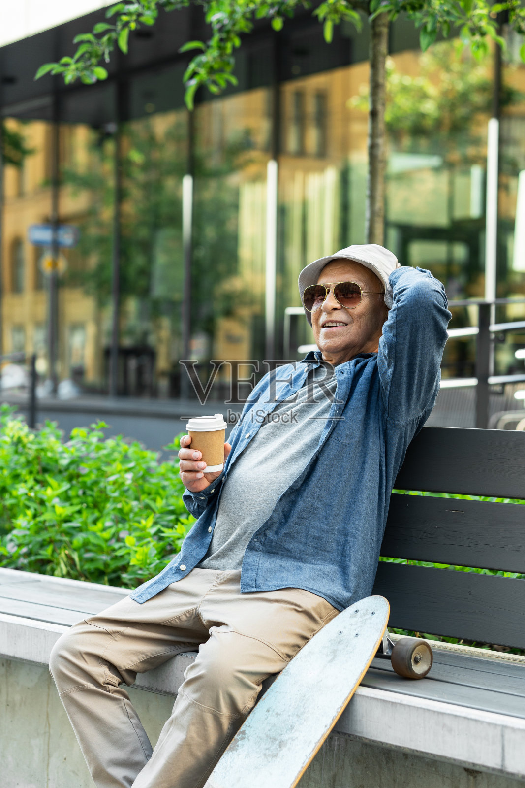 在城市公园里，一个拿着长板的老人坐在长凳上喝咖啡照片摄影图片