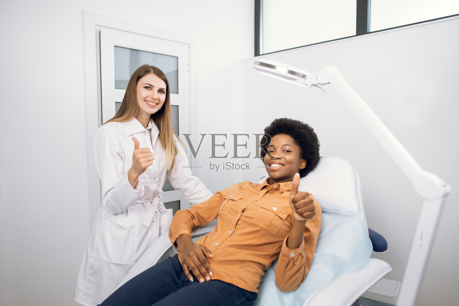 快乐年轻的非洲裔美国人微笑的女人，满意的结果美容医学诊所的美容程序，显示拇指与她的女医生美容师相机。照片摄影图片