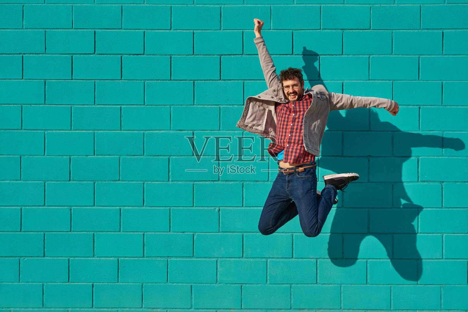 正面的观点，一个快乐的人跳上一个蓝色明亮的墙在一个阳光明媚的日子照片摄影图片