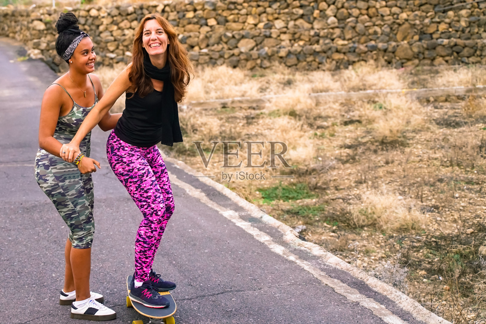年轻的母亲在农村的街道上练习滑板。女儿帮妈妈玩滑板。妈妈学习骑滑板，女儿教她在郊区的街道有乐趣-形象照片摄影图片