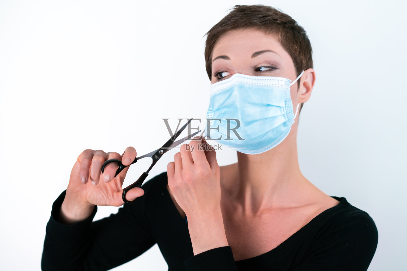漂亮的短发女人用剪刀摘除她的面具。生活在隔离。应对冠状病毒限制。生活在大流行。结束封锁。照片摄影图片