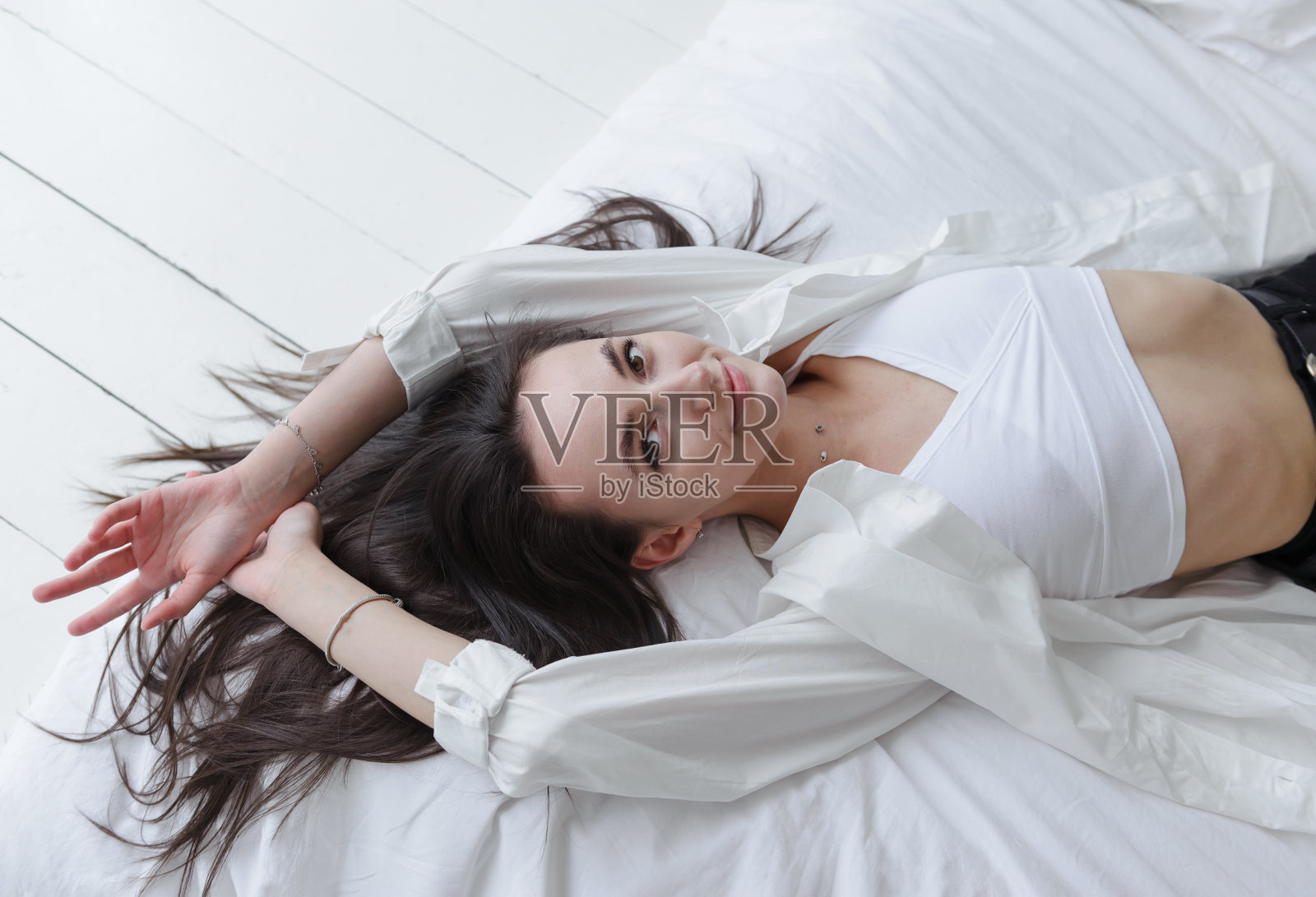 白色年轻漂亮的黑发女子，穿着白色衬衫拿着手机，长发躺在床上笑着照片摄影图片