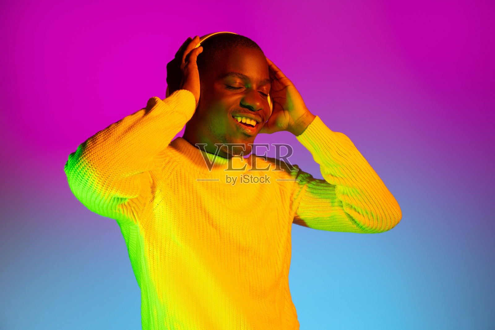 年轻快乐的非洲英俊男子与耳机隔离在梯度品红色黄色霓虹灯背景。照片摄影图片