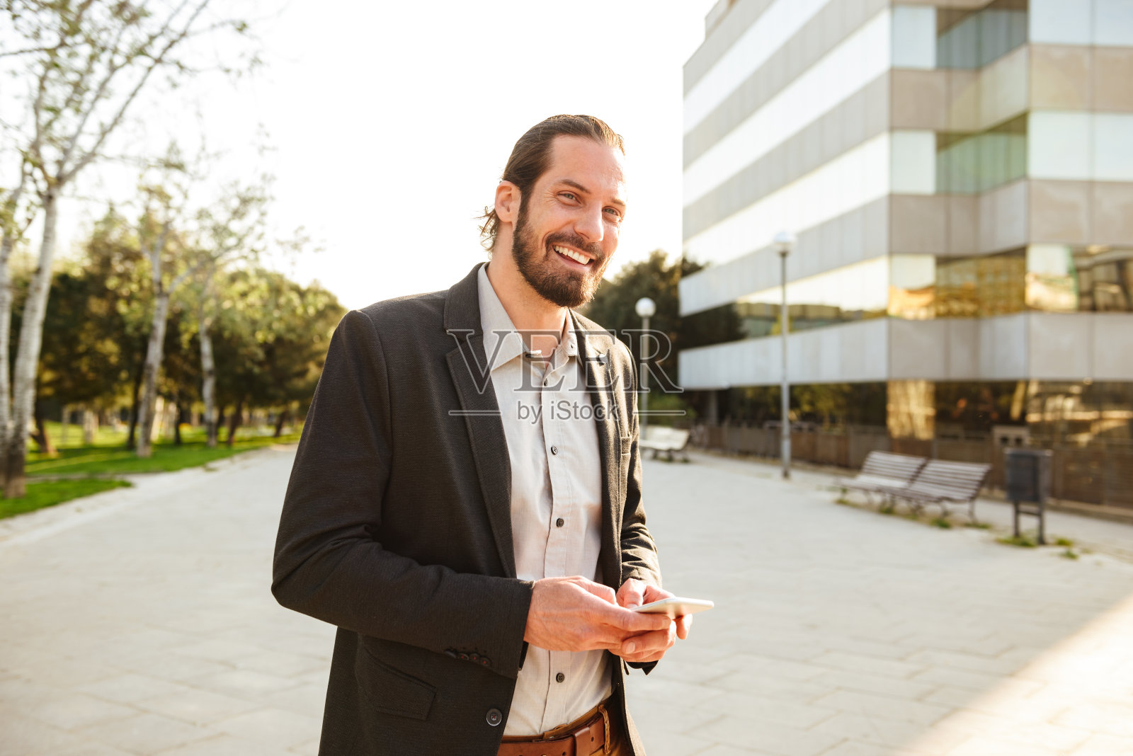 照片中的白人微笑的男人30多岁的商务西装看着你和拿着智能手机，而走在办公大楼附近照片摄影图片