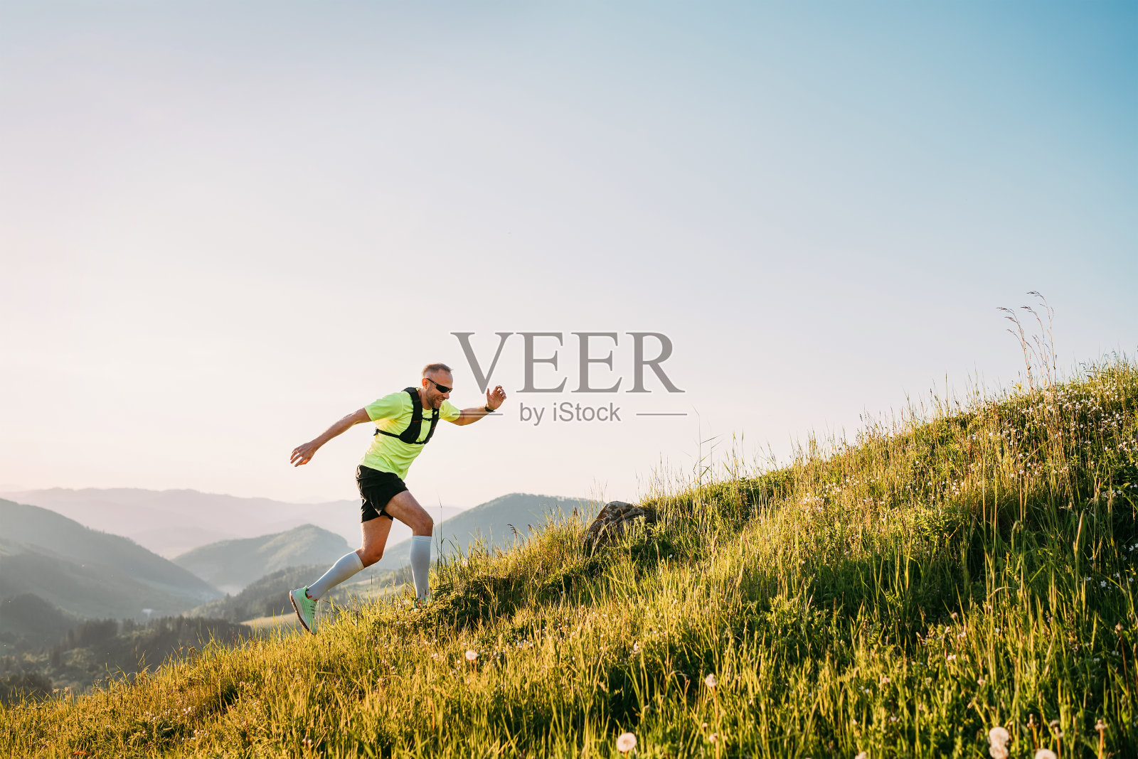 中年的登山运动员穿着明亮的t恤，背着运动太阳镜的背包，在日落时分沿着风景如画的山丘往山上跑。运动积极的人概念形象。照片摄影图片