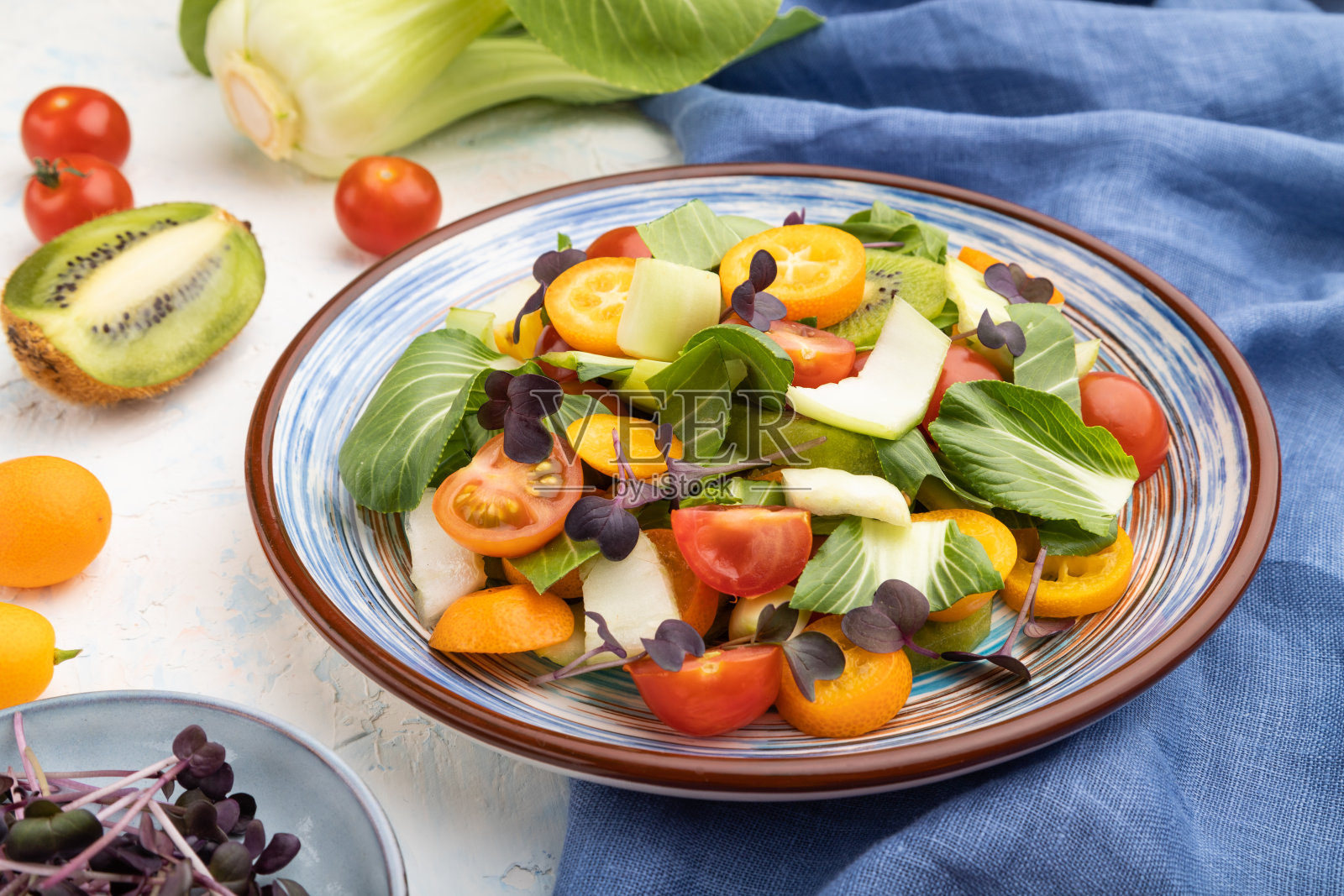 素菜沙拉，卷心菜，猕猴桃，番茄，金橘，微绿芽，白色混凝土背景和蓝色亚麻织物。侧视图，特写。照片摄影图片