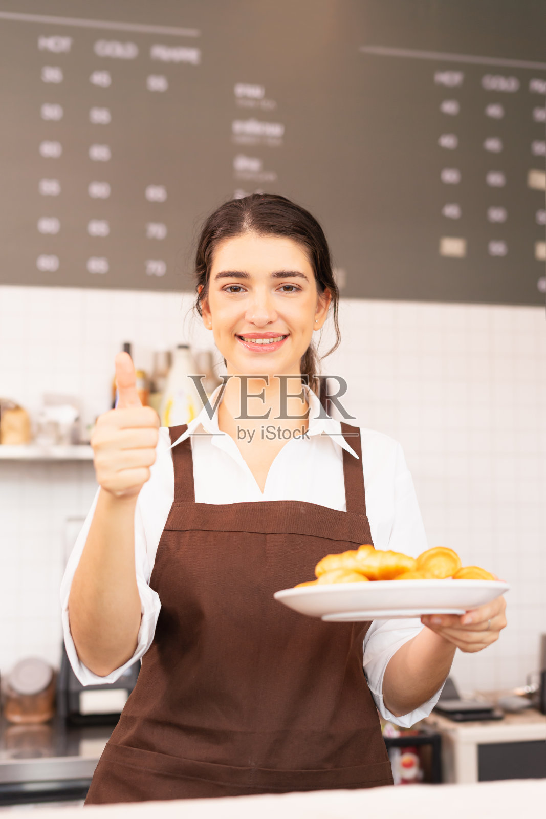 垂直框架的美丽的白人咖啡师女人看左边，拿着盘子与面包在手上，并给予拇指在咖啡吧。照片摄影图片