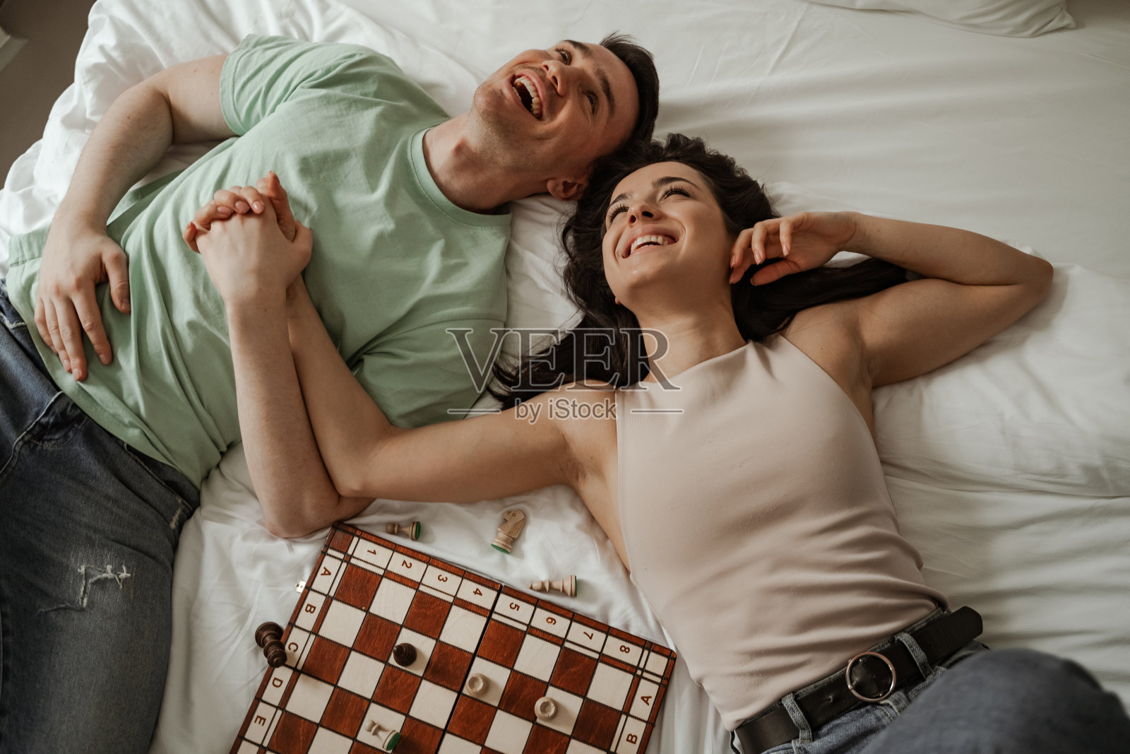 幸福的年轻夫妇躺在床上接吻，床上放着象棋。年轻的家庭搬到新的公寓照片摄影图片