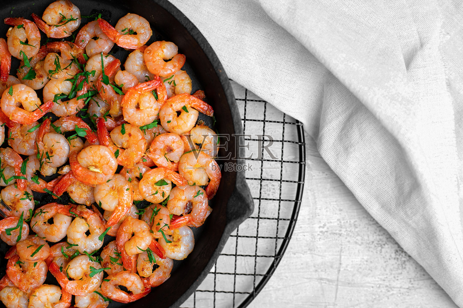 在白色的餐桌上，放着白色的亚麻布餐巾，在铁锅里与大蒜一起煎的大虾。前视图。照片摄影图片