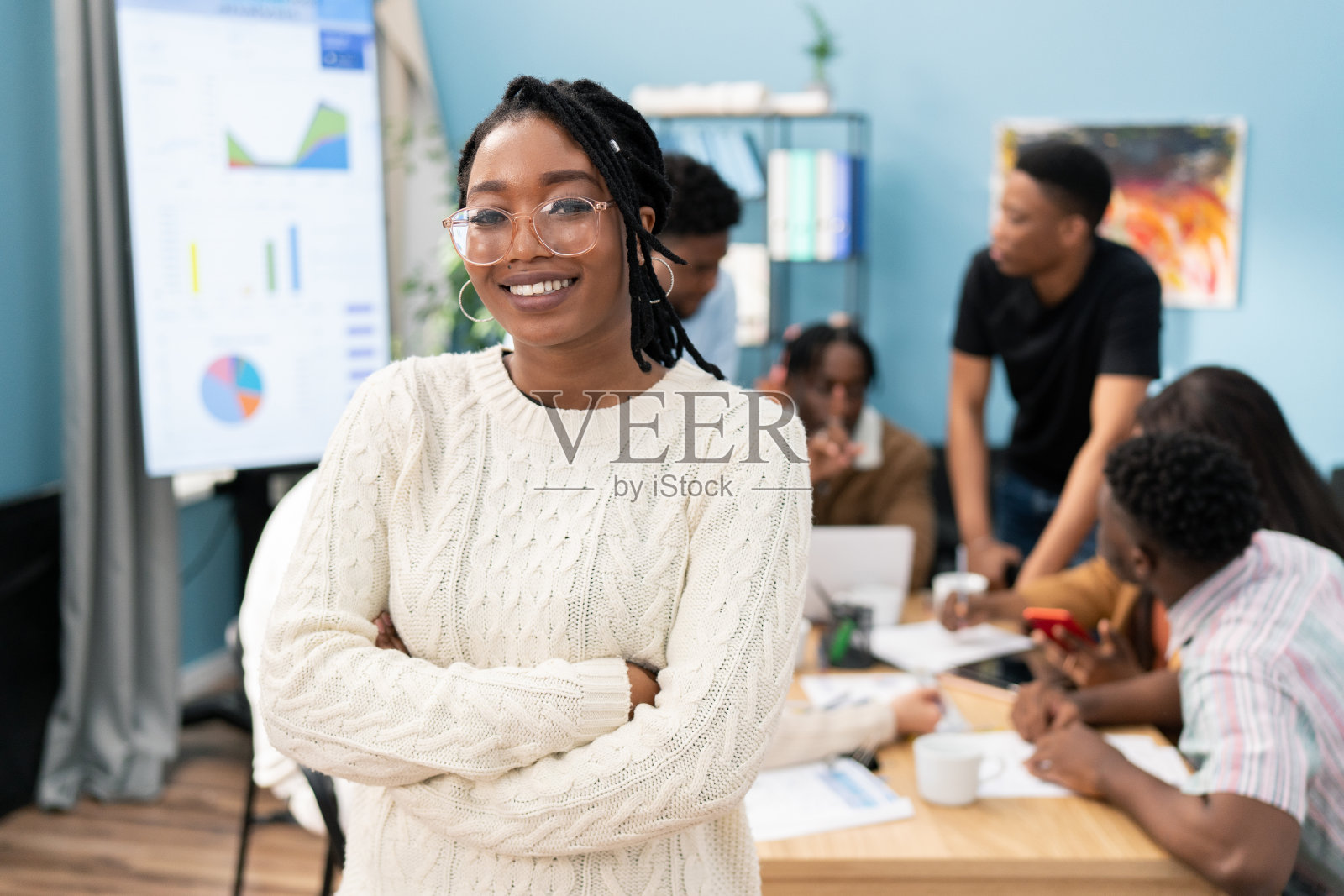 一个微笑的皮肤黝黑的女孩穿着一件暖和的毛衣，双臂交叉在胸前，女人戴着眼镜和金耳环，她的头发编成辫子，背景是一个商务会议在办公室照片摄影图片