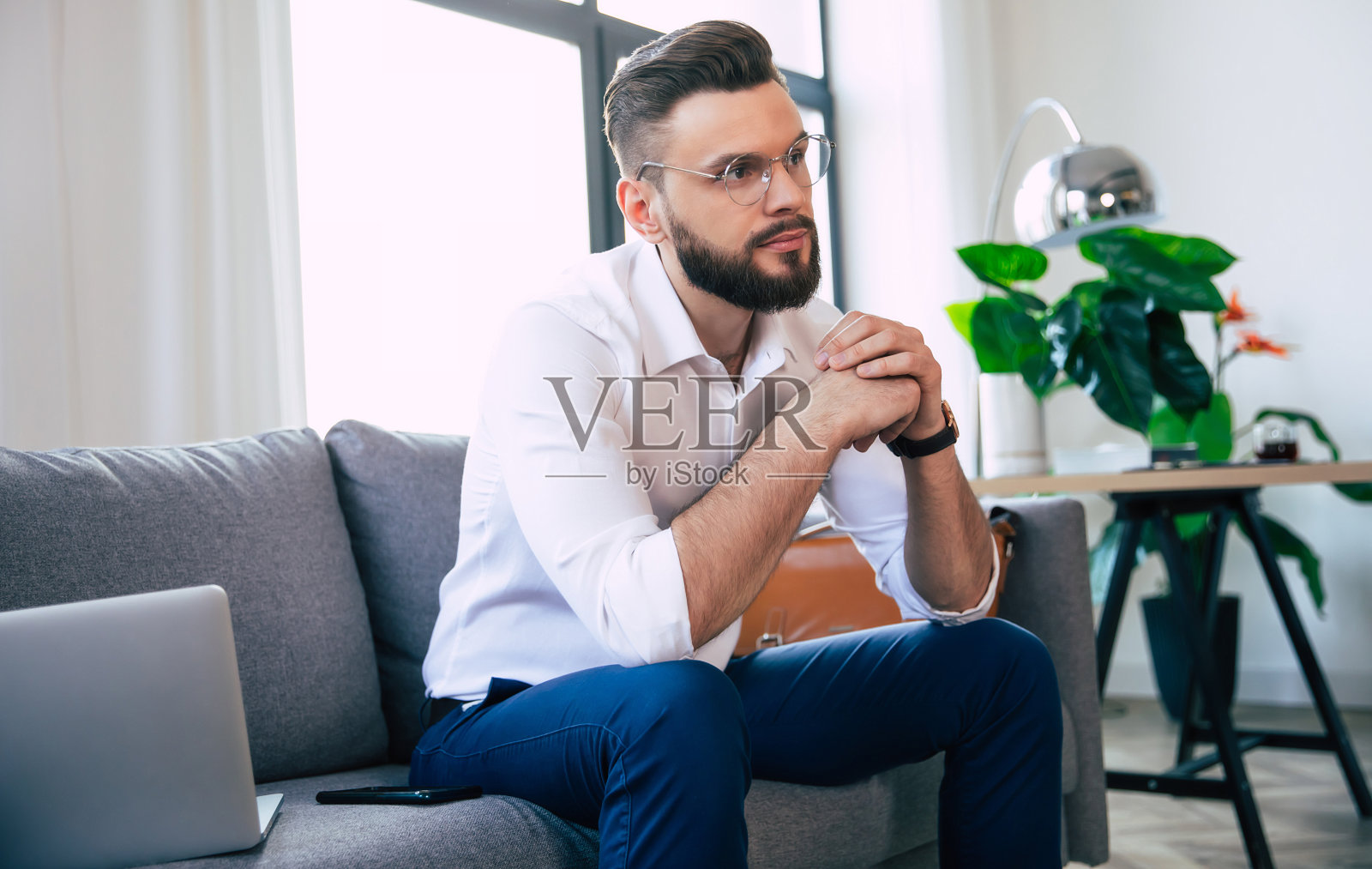 自信、时尚、蓄着胡须、戴着眼镜、穿着正装的商务男士正坐在现代办公室或房间的沙发上照片摄影图片