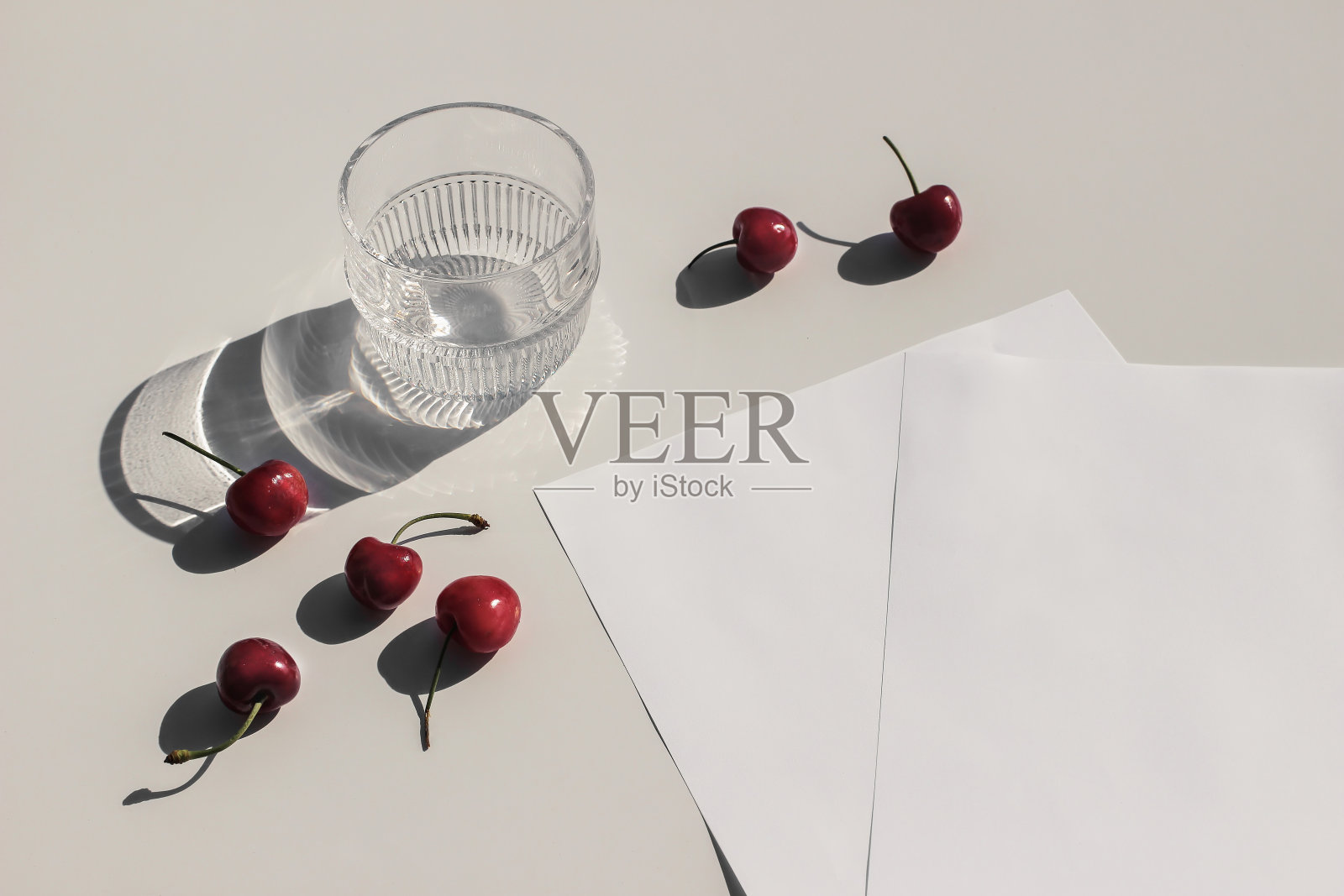 夏季食物静物组成。米色餐桌背景上的鲜红樱桃。文具模拟场景。白纸页面。一杯水，一杯在阳光下闪闪发光的鸡尾酒。前视图。照片摄影图片