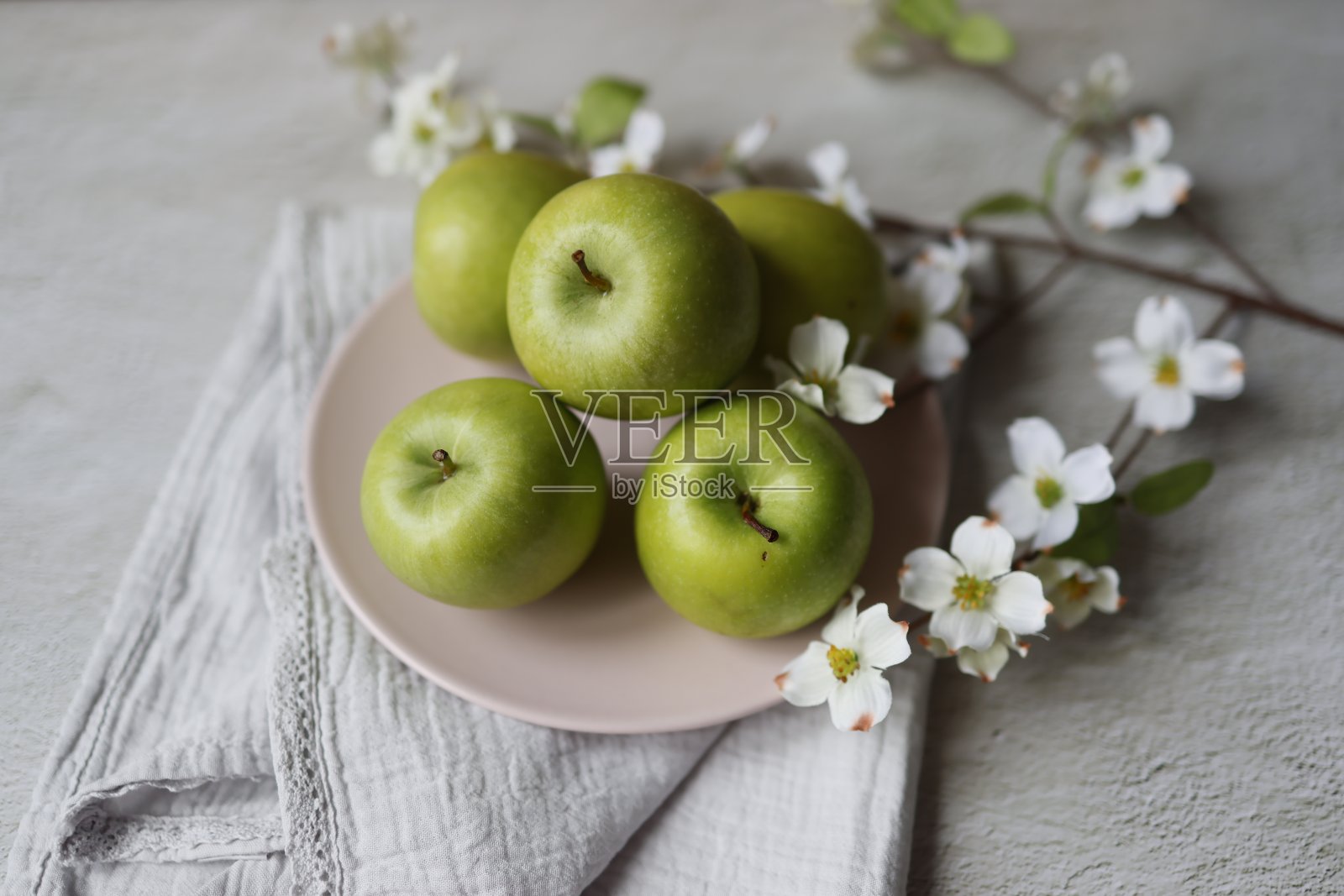 浅灰色粗糙的混凝土地板上，浅粉色的盘子上放着青苹果，盘子周围是开着白花的山茱萸照片摄影图片