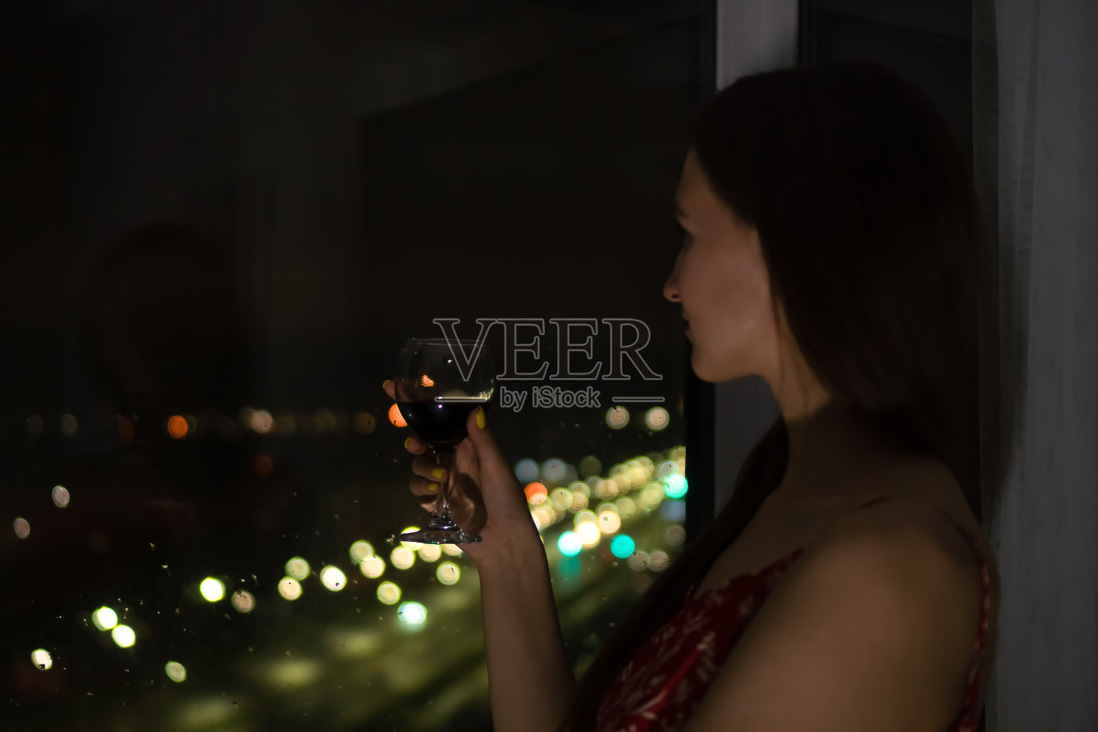 年轻的黑头发的女人，身穿红色的带子和一杯红酒站在窗边，看着城市的夜生活。有选择性的重点照片摄影图片