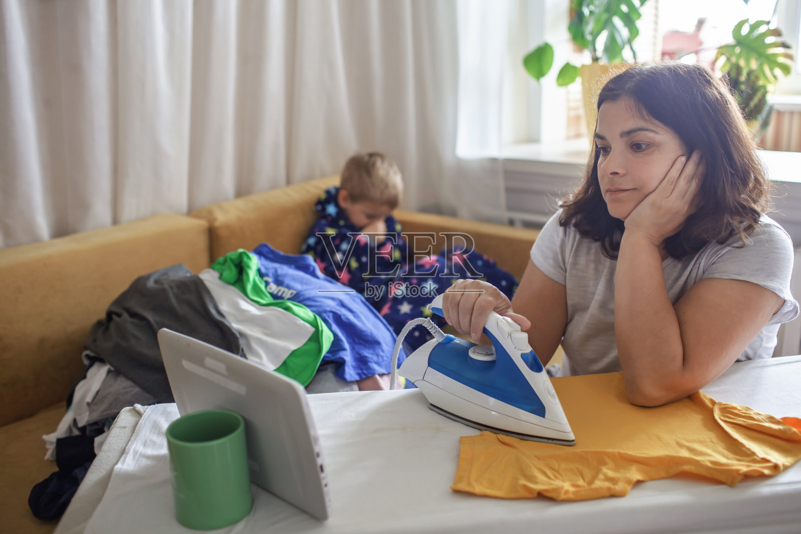 疲惫的妈妈边看平板视频边熨烫衣物，儿子在旁边玩手机照片摄影图片