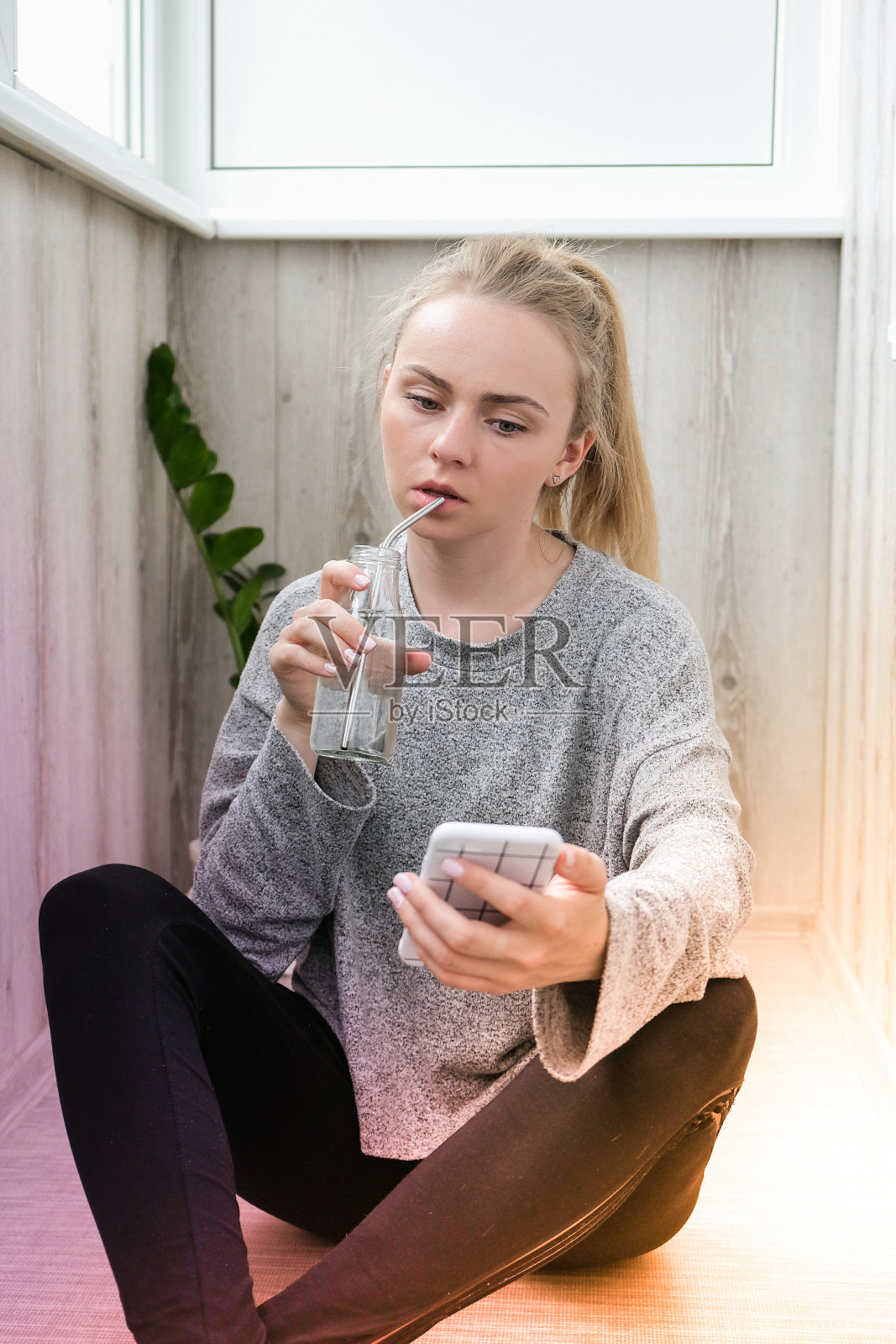 年轻的千禧金发女人喝着环保瓶子里的水和金属可重复使用的吸管，在阳台上做瑜伽锻炼伸展健身。正念冥想。健康营养、环保、零浪费的理念照片摄影图片