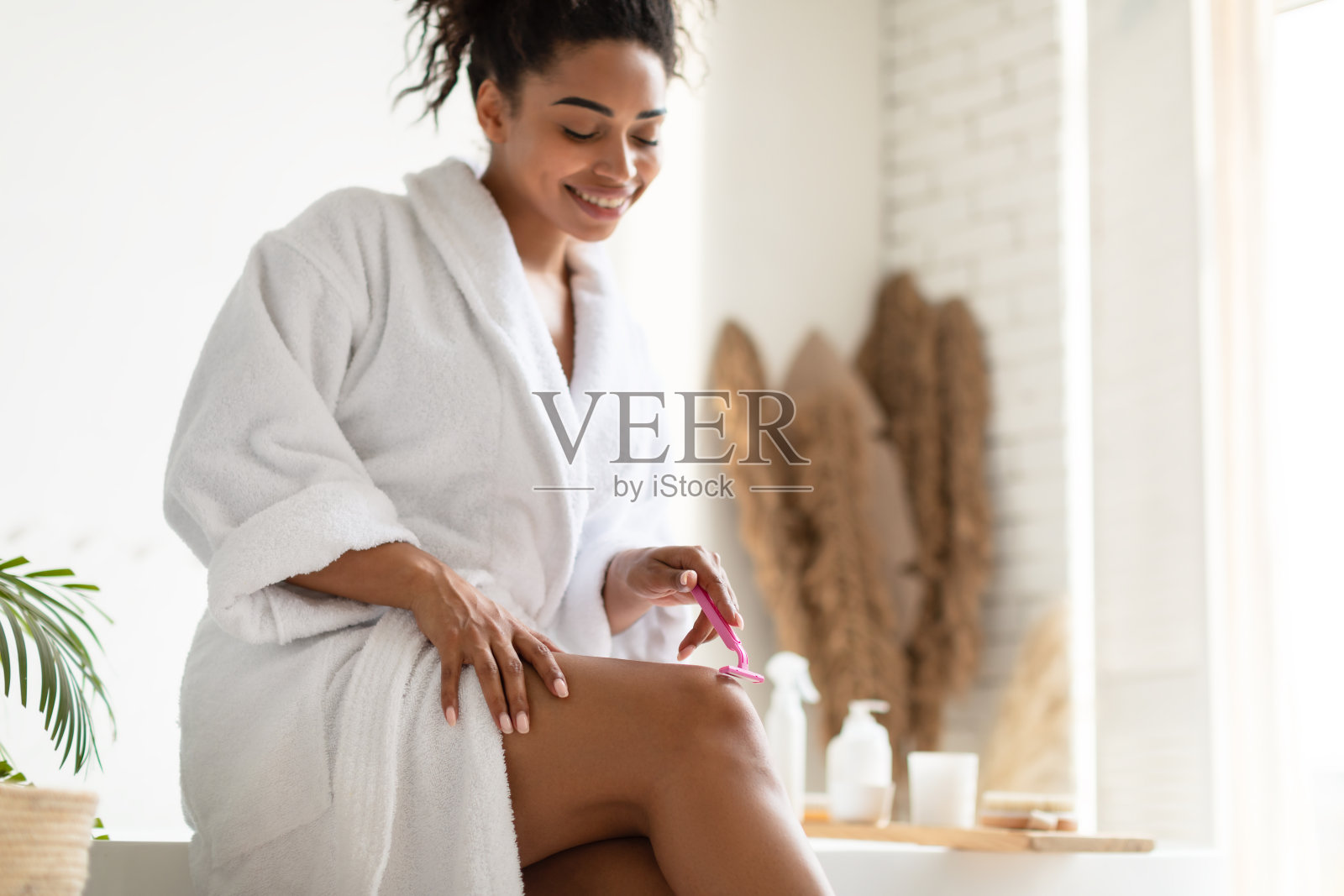 快乐的黑人妇女使脱毛剃腿在现代浴室照片摄影图片